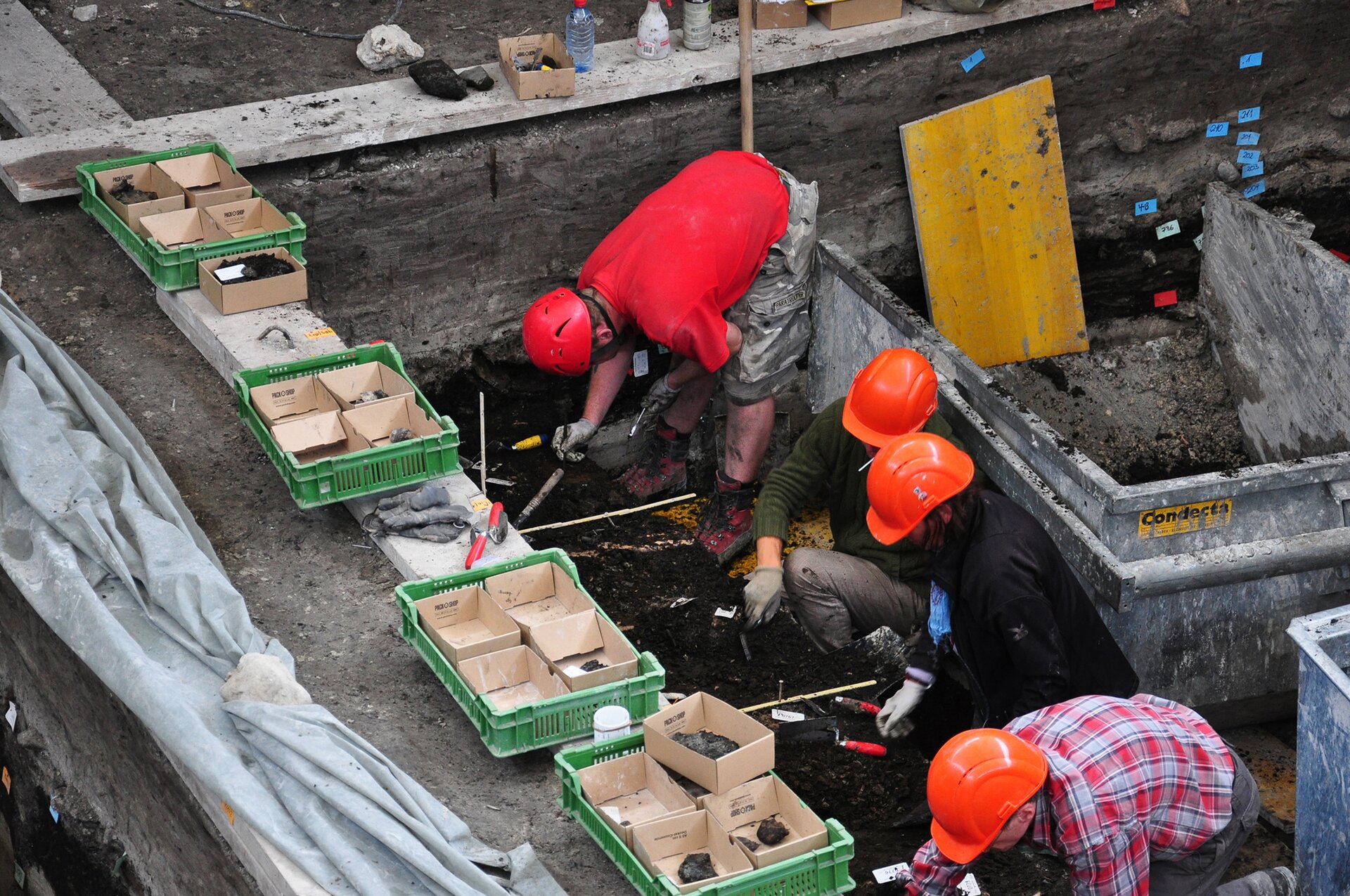 Zdjęcie przedstawia czterech archeologów przy pracy. Na głowach mają kaski. Kucają i pochylają się i grzebią w ziemi. Przed nimi stoją plastikowe kosze z kartonowymi pudełkami, gdzie posegregowane są archeologiczne skarby. Za nimi stoją kontenery ze żwirem i piaskiem. 