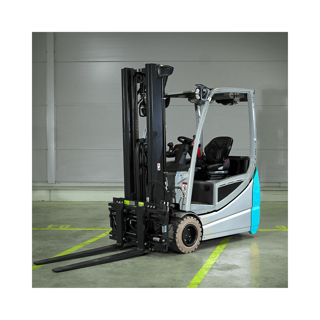 Zdjęcie przedstawia wózek widłowy. Jest to mały pojazd z miejscem dla jednej osoby. Z przodu wózka znajduje się widelec o dwóch ostrzach mogący poruszać się w górę i w dół po dwóch pionowych szynach.