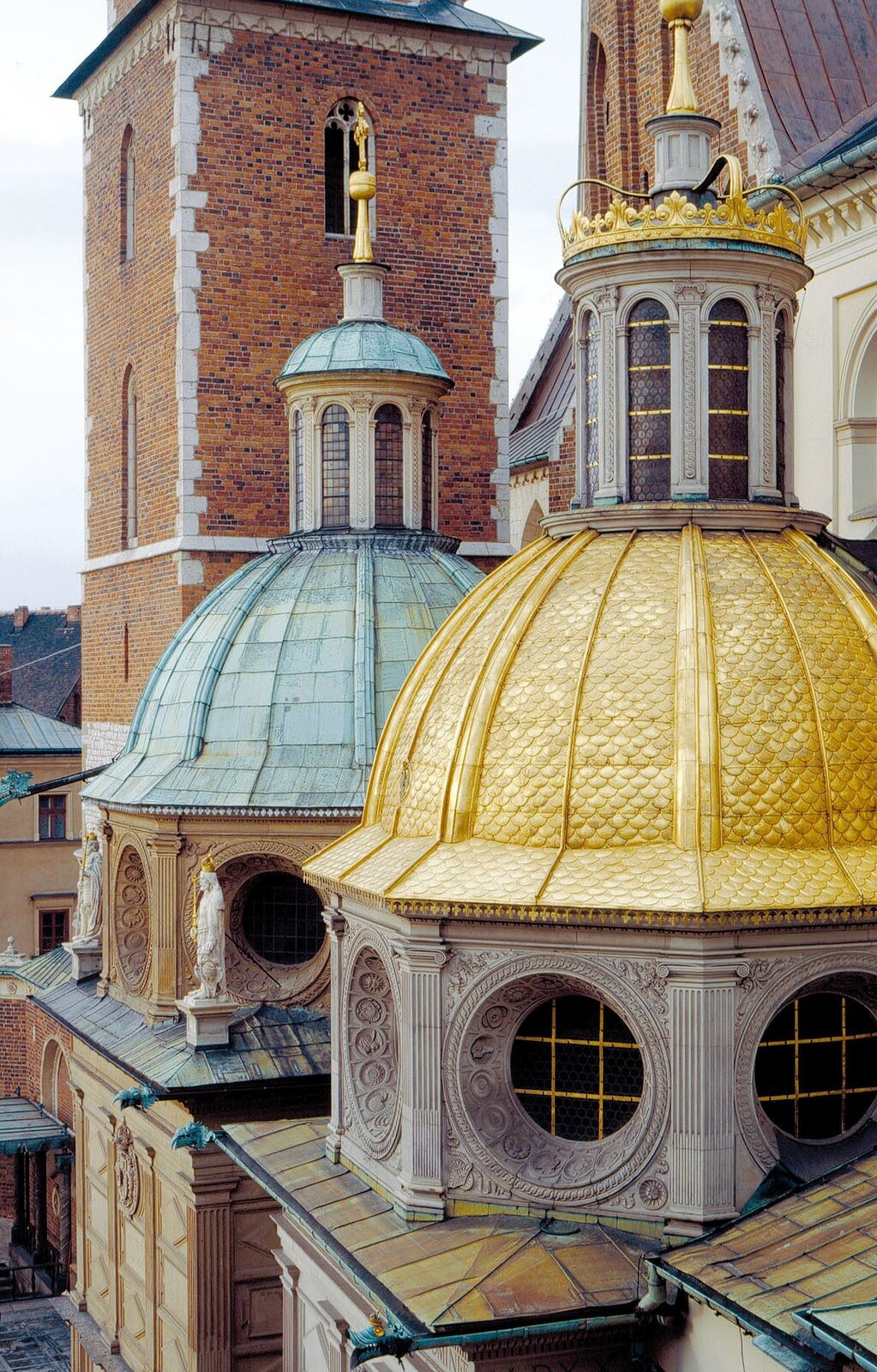 Ilustracja przedstawia zbliżenie na złotą kopułę kaplicy Zygmuntowskiej. Za nią znajduje się kaplica Wazów, w tle - wieża kościelna tzw. Srebrnych Dzwonów, zbudowana z czerwonej cegły.