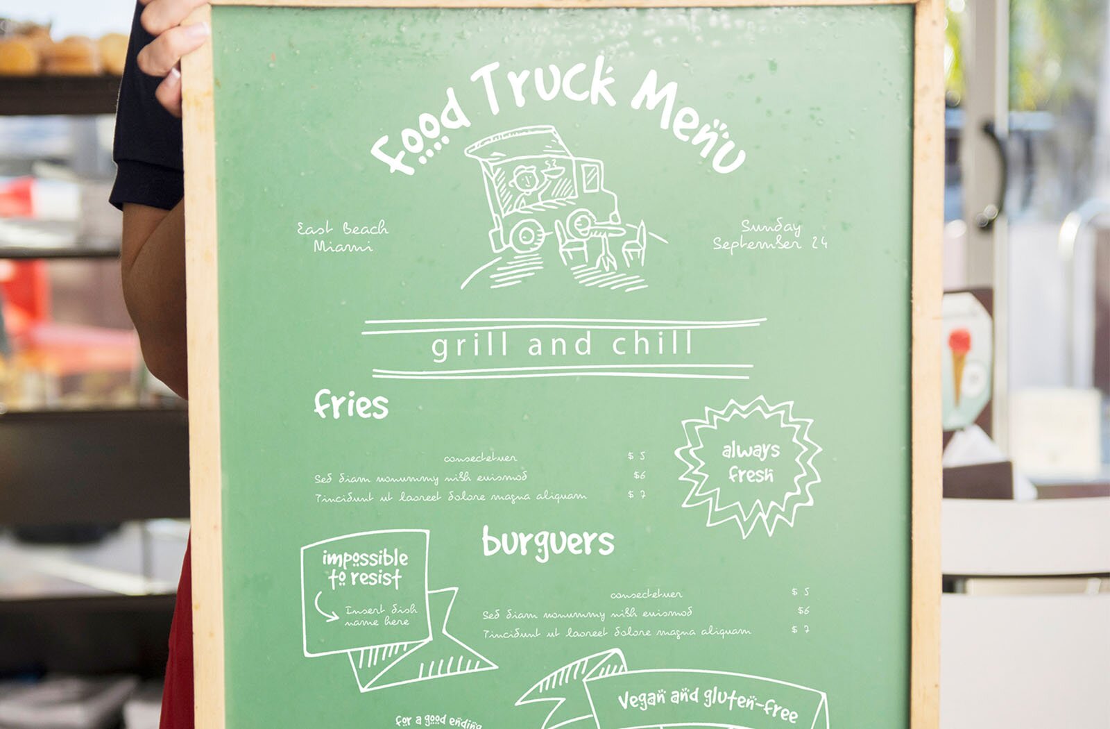 Zdjęcie przedstawia zieloną tablicę w drewnianej ramie. Na tablicy widoczne są białe napisy w języku angielskim. Na górze widoczny jest napis: Food Truck Menu, pod którym widać rysunek food trucka. Po jego lewej stronie jest napis: East Beach Miami, a po prawej – Sunday September 24. Poniżej między podwójnymi liniami widać napis: grill and chill. Następnie widoczne jest menu z cenami oraz napisy: fries, always fresh, burgers, impossible to resist, vegan and glute‑free.