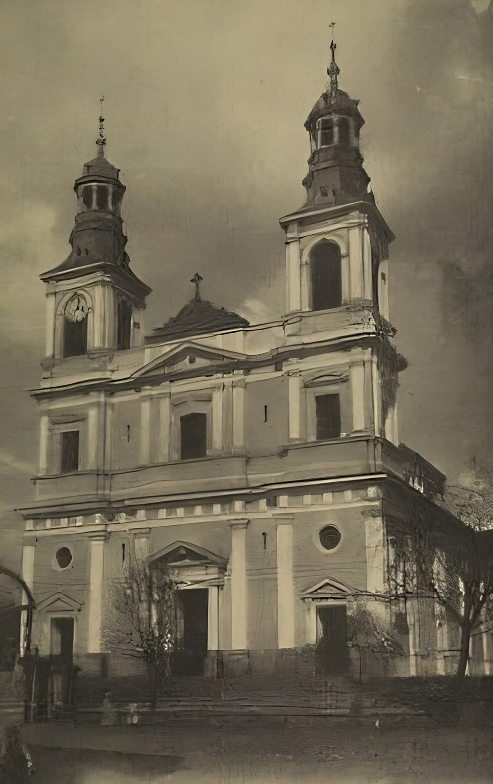 Kliknij, aby powiększyć Zdjęcie przedstawia neobarokową świątynię z dwoma wieżami, nakrytymi hełmami z sygnaturką i ozdobione dużymi oknami. Fasada wejściowa jest dwukondygnacyjna podzielona pilastrami i wydzielona gzymsem z balustradą. Wejście jest pod trójkątnym tympanonem ocienionym dwoma kolumnami. Podobny tympanon z czterema kolumnami po dwie po obu stronach okna balkonowego znajduje się na piętrze kościoła. Wejścia do naw bocznych na parterze również naśladują wejście główne.