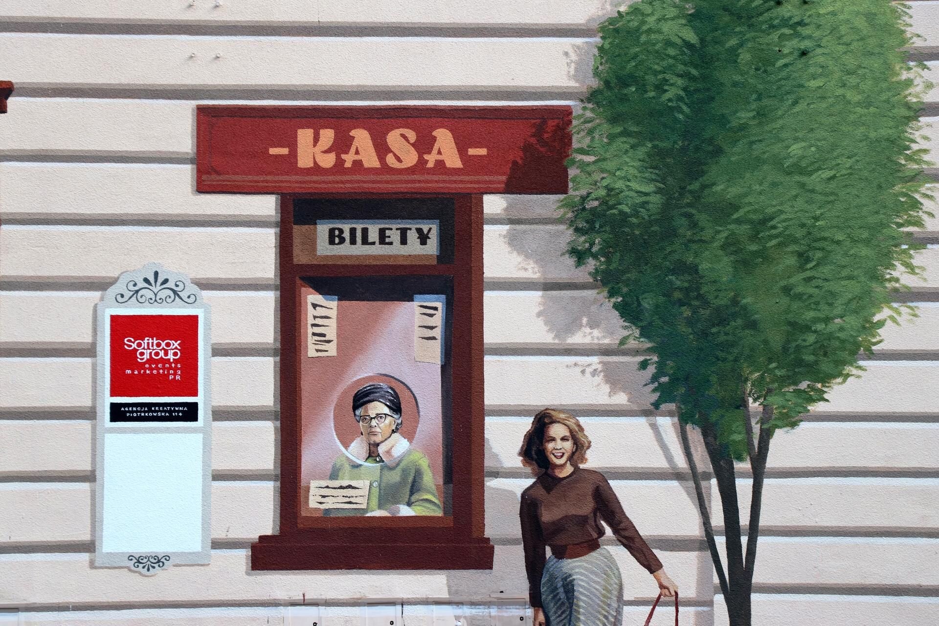 Zdjęcie przedstawia mural, czyli obraz na fasadzie budynku. Praca przedstawia białą ścianę ze żłobieniami, w centrum ściany widoczna jest starsza kobieta ubrana w zielony płaszcz, która siedzi w okienku biletowym. Nad okienkiem ciemno‑czerwona tabliczka z napisem: Kasa. Z prawej strony okienka kasowego rośnie zielone liściaste drzewo, obok którego stoi młoda kobieta z jasnymi średniej długości włosami, ubrana w brązową bluzkę i pasiastą sukienkę. Z lewej strony okienka kasowego wisi prosty plakat o czerwonym tle, na którym widnieją białe napisy: Softbox group [czytaj: softboks grup].