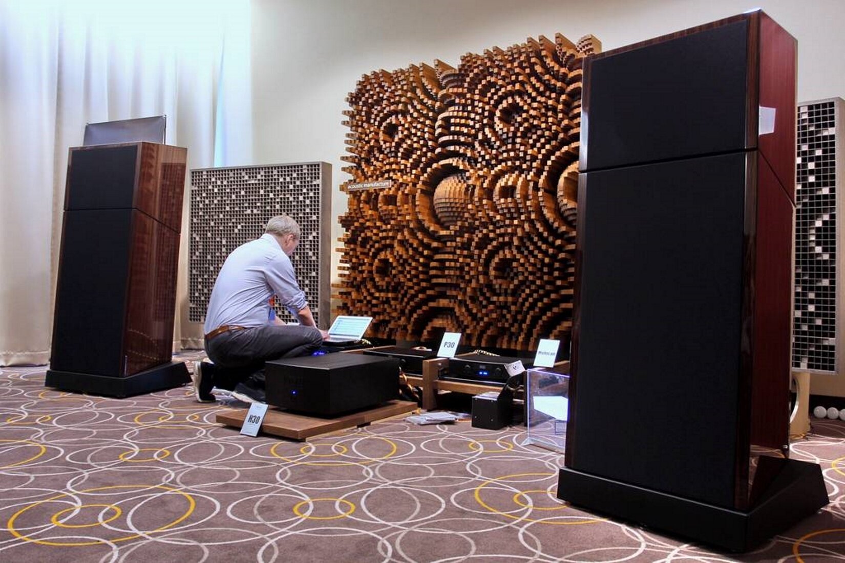 Zdjęcie przedstawia oparty o szarą ścianę dyfuzor drewniany w kształcie kwadratu.  Składa się z wypukłych, poziomych kolców. Całość konstrukcji tworzy okrągłe pętle, które nachodzą na siebie. W jasnym pomieszczeniu przed dyfuzorem na szarym dywanie w okrągłe biało‑żółte pętle znajduje się klęczący tyłem mężczyzna, który ma przed sobą otwartego laptopa z ułożonymi na nim dłońmi. Ma na sobie jasnoszarą koszulę i długie szare spodnie z brązowym paskiem oraz czarne buty.  Po prawej stronie mężczyzny znajdują się położone na drewnianych deskach odtwarzacze, a po prawej i lewej stronie ilustracji znajdują się dwa, wysokie, drewniane głośniki. Na ścianie po prawej i lewej stronie dyfuzora znajdują się dwie metalowe, kraty. 