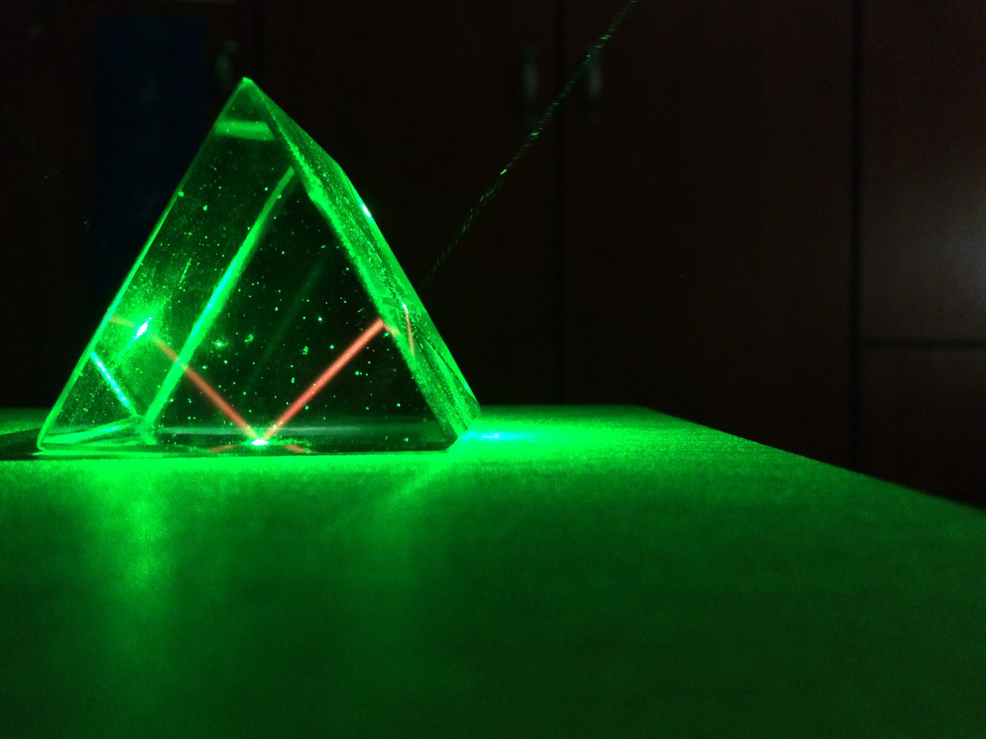 Zdjęcie przedstawia pryzmat trójkątny, przez który przechodzi promień z lasera. Pryzmat leży na stole, w ciemnym pokoju. Wokół pryzmatu zielona poświata. Od lewej strony wpada do niego wiązka laserowa, pod takim kątem, że następnie pada na podstawę pryzmatu, odbija się pod takim samym kątem, i wychodzi z pryzmatu. W pryzmacie wiązka jest czerwona; poza nim, na jej drodze widać świecące na zielono drobiny kurzu.
