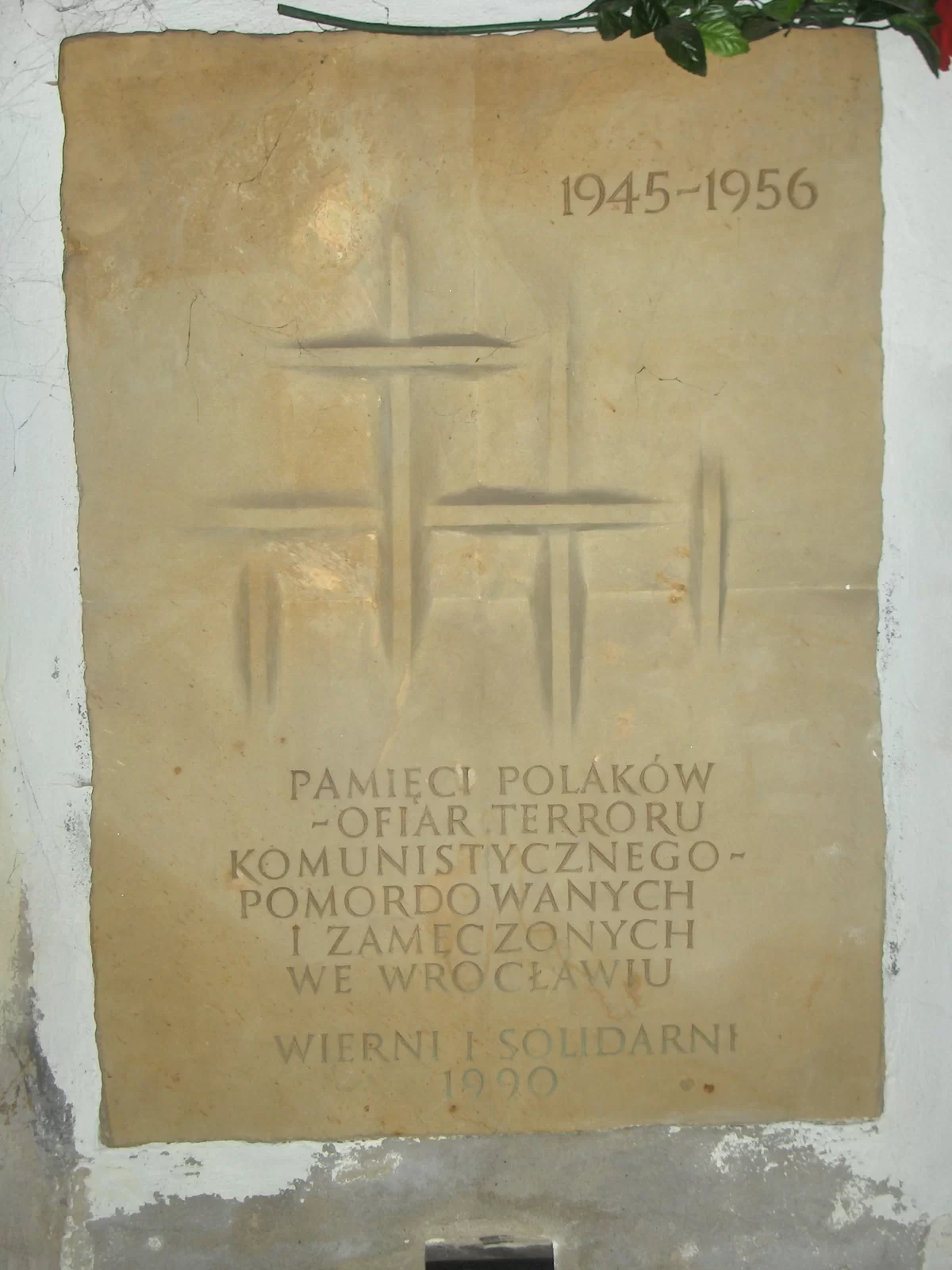 Kliknij żeby powiększyć Kamienna tablica wisi na ścianie domu. Na tablicy znajdują się: po prawej stronie u góry daty (1945‑1956), po środku wyryte są krzyże, a na samym dole napis: Pamięci Polaków - ofiar terroru komunistycznego pomordowanych i zamęczonych we Wrocławiu. Wierni i solidarni 1990.