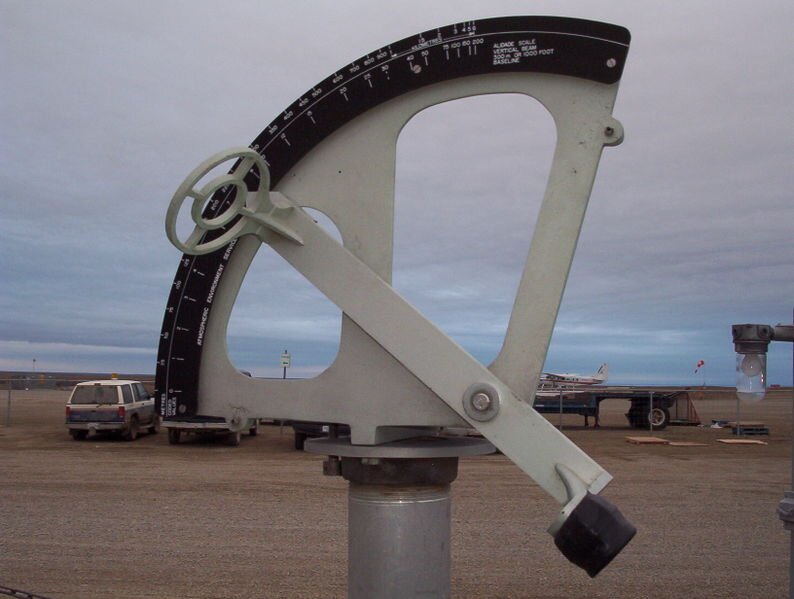 Na zdjęciu znajduje się alidada. Ma kształt wyciętej z okręgu części. Dolna część jest zaostrzona, boki ustawione są prawie pod kątem prostym, natomiast górna część jest zaokrąglona. Na górnej części znajduje się miarka. Przez alidadę przechodzi metalowy, płaski element, który ma na obu końcach wypukłe elementy. Alidada ustawiona jest na metalowym pręcie. 