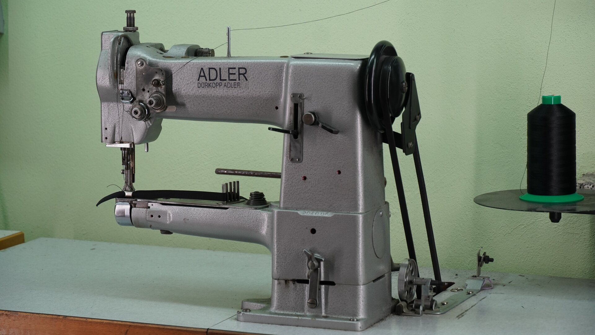 Zdjęcie przedstawia szarą maszynę - lamowarkę. Z jej boku znajduje się pokrętło, od którego odchodzą dwie taśmy. Znikają one w otworze blatu, na którym stoi urządzenie. Nagranie tożsame z treścią.