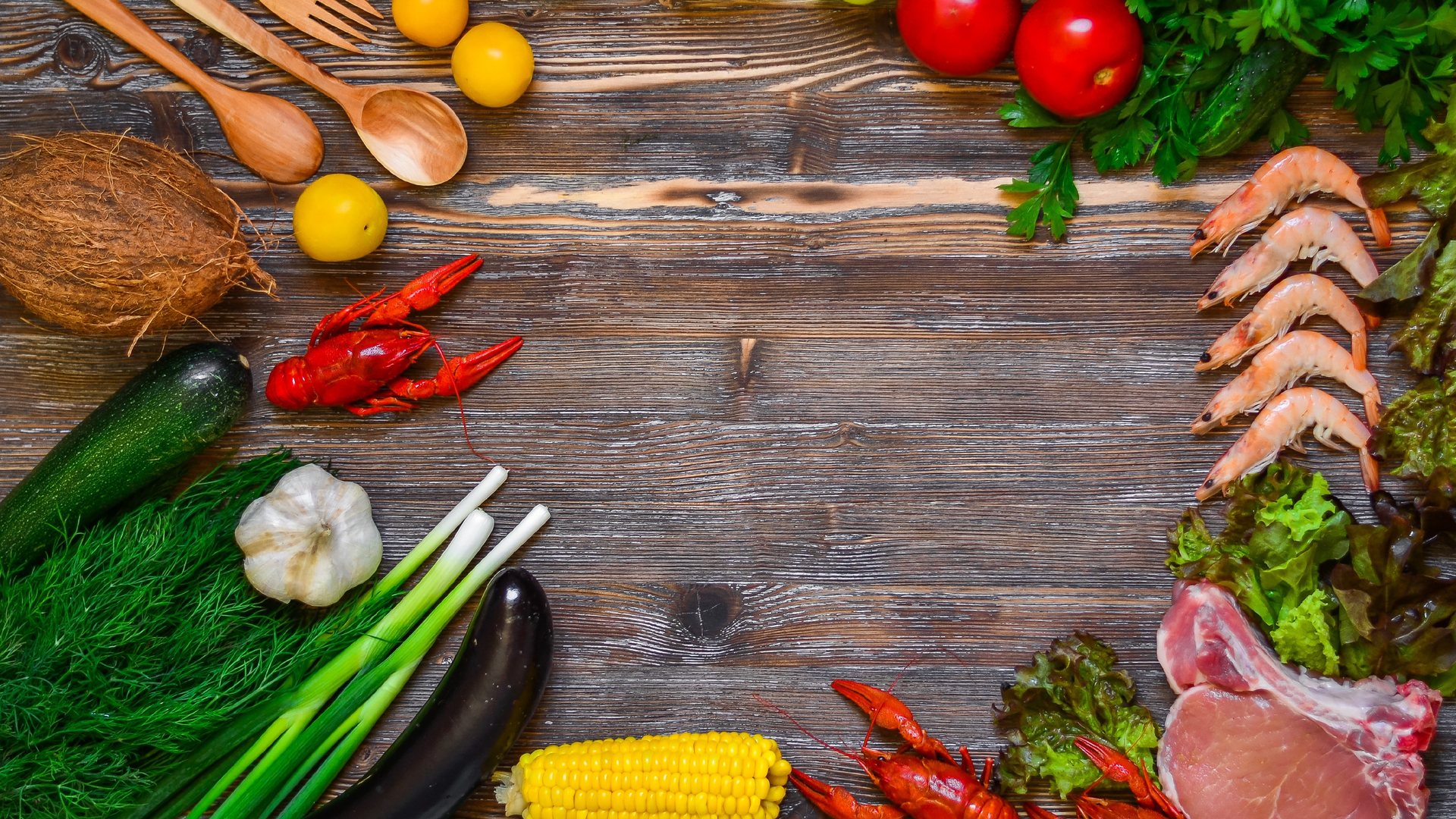 Zdjęcie wykonane z góry przedstawia produkty żywnościowe, które ułożone są na blacie z desek. Po lewej stronie na zdjęciu znajdują się warzywa: cukinia, bakłażan, kolba kukurydzy, dymka, główka czosnku oraz koperek. Po lewej stronie u góry na zdjęciu znajduje się kokos, mały krab, żółte pomidory. Obok leżą drewniane łyżki oraz widelec. Po prawej stronie ilustracji widoczne są: dwa pomidory, kilka krewetek, plaster surowego mięsa oraz natka i liście sałaty.