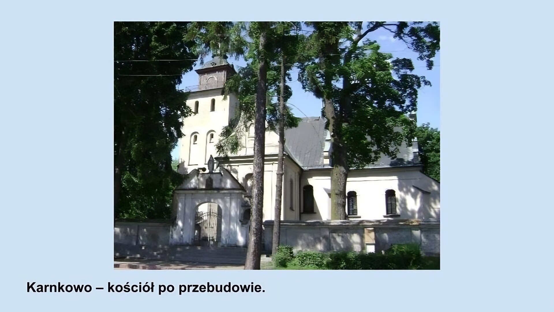 Ilustracja przedstawia kościół w Karnkowie po przebudowie. Biały budynek ze spadzistym szarym dachem, oknami z łukami, wieżą po środku. Przed budynkiem widać drzewa oraz biały mur z bramą z figurką matki boskiej na jej szczycie.