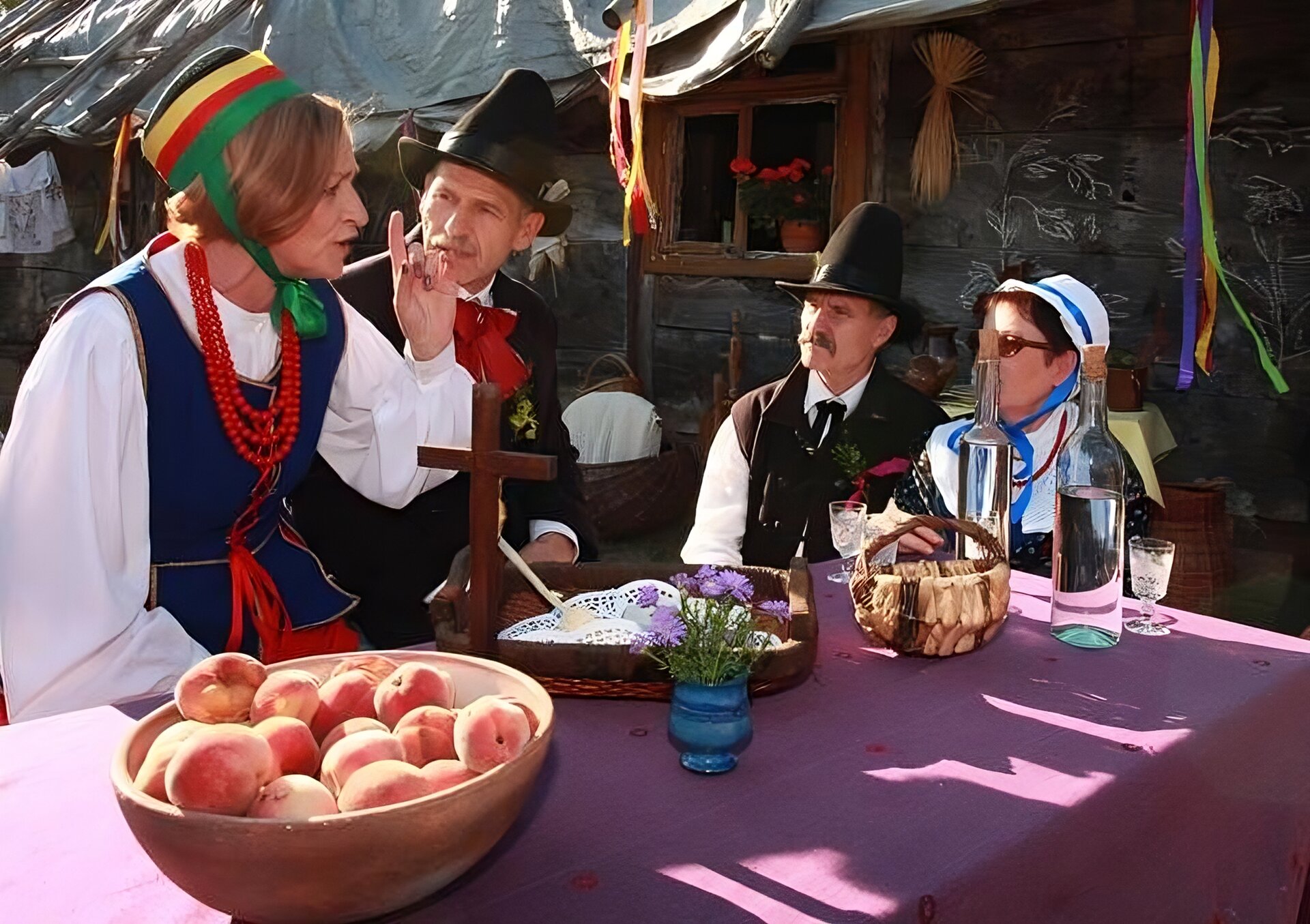 Zdjęcie przedstawia cztery osoby ubrane w stroje ludowe. Siedzą one przy stole nakrytym fioletowym materiałem, na którym stoi drewniany krzyż, dwie drewniane misy, w której w jednej z nich znajdują się brzoskwinie, niebieski wazonik z fioletowymi kwiatami, dwie szklane butelki wypełnione bezbarwnym płynem oraz wiklinowy kosz. Pierwsza osoba z lewej strony zdjęcia jest kobietą, która ubrana jest w białą koszulę, niebieską kamizelkę, czerwone korale, zielono‑czerwono‑żółto‑czarną czapkę. Kobieta podnosi palec wskazujący do góry. Obok niej siedzi mężczyzna w czarnej marynarce, białej koszuli, czerwonej kokardzie przypiętej do szyi oraz czarnym kapeluszu. Patrzy się w kierunku mówiącej kobiety. Obok niego siedzi drugi mężczyzna ubrany do niego podobnie - ma on czarną, a nie czerwoną kokardę. Ma wąsy i wpatruje się w kobietę. Obok niego siedzi druga kobieta ubrana w czarną suknię z białym, dużym kołnierzem, czerwone korale i biało‑niebieską czapkę. Ona również wpatruje się w mówiącą kobietę. W tle widoczny jest fragment starego budynku. Zdjęcie zostało wykonane w świetle dziennym.