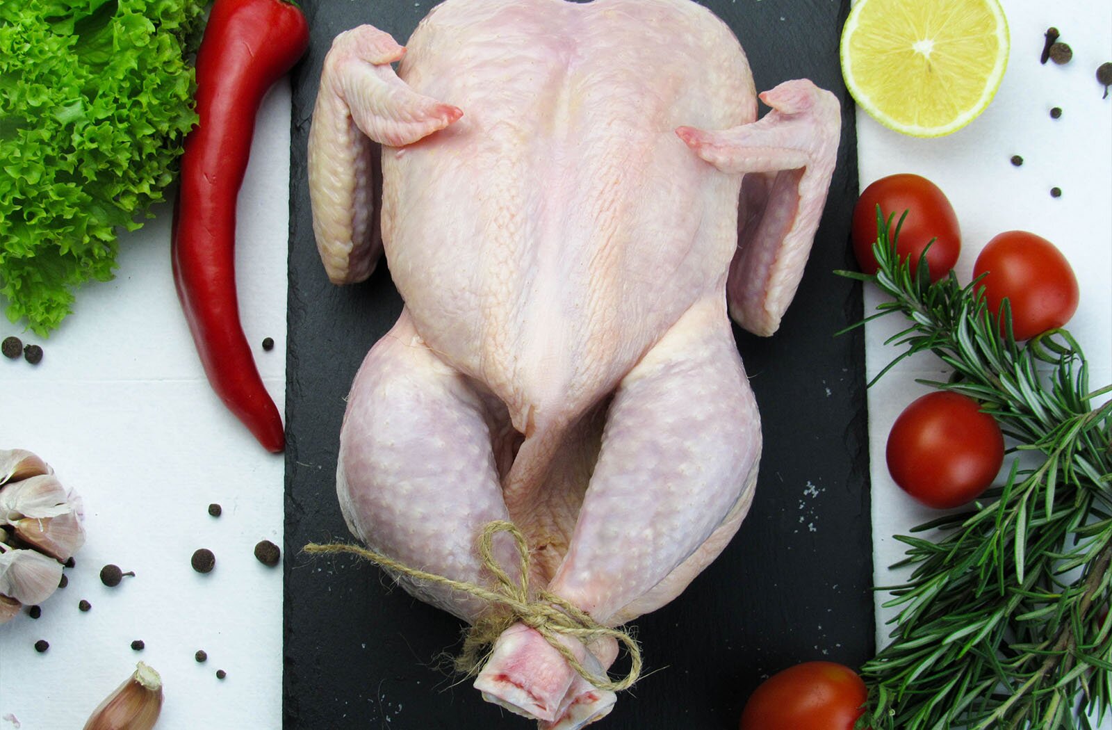 Zdjęcie przedstawia indyka przygotowanego do pieczenia, ułożonego na czarnej desce. Indyk ma związane sznurkiem nogi. Po prawej i lewej stronie deski na blacie widoczne są małe pomidorki, połówka cytryny, ostra czerwona papryka, gałązki rozmarynu, liście karbowanej zielonej sałaty, główka czosnku i ziarna czarnego pieprzu.