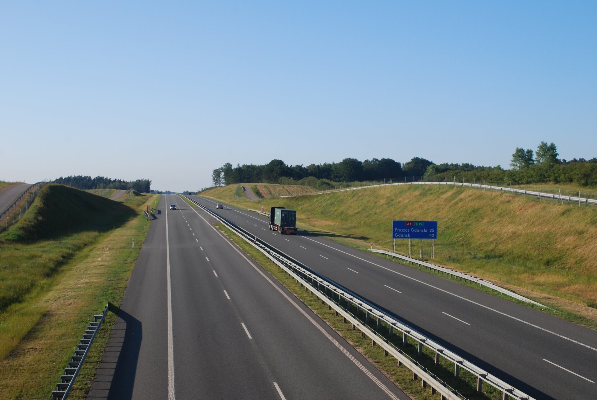 Na zdjęciu prosty odcinek autostrady, dwa pasy drogi i pobocze w każdym kierunku, ogrodzona siatką.
