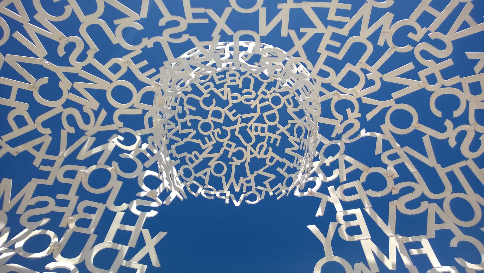 Kolorowe zdjęcie rzeźby wykonane z jej wnętrza, przedstawia litery połączone ze sobą na tle nieba. W centrum litery układają się w okrągłą wypukłość.