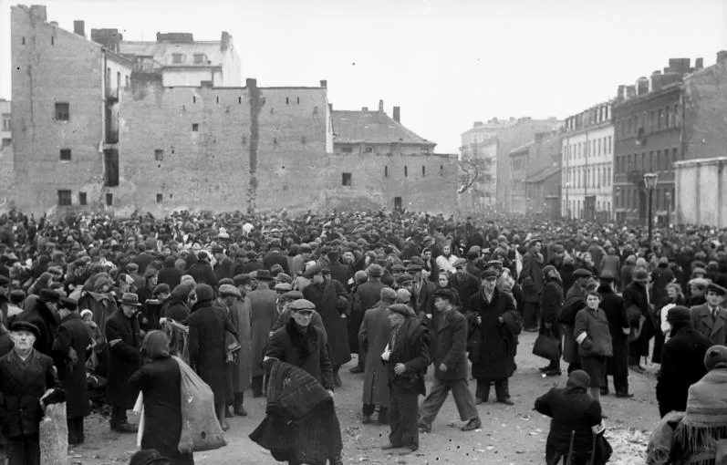Fotografia przedstawia duży plac. Chodzi tu tłum ludzi. Wielu z nich nosi na plecach worki lub płaszcze przewieszone przez ramiona. Są ubrani w długie płaszcze lub kurtki. Na prawych ramionach mają opaski. W tle stoją zniszczone, kilkukondygnacyjne kamienice.