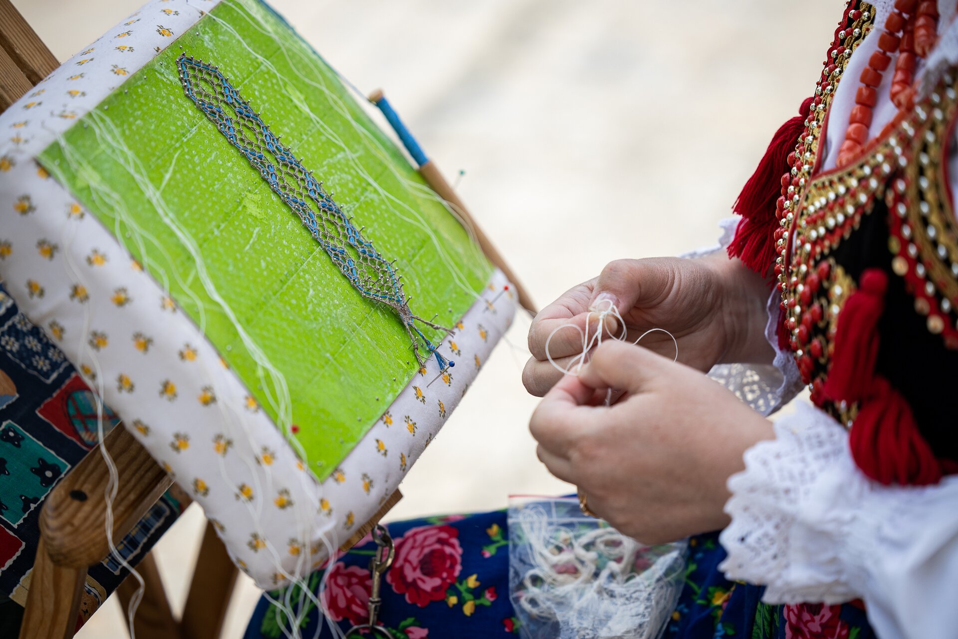 Zdjęcie przedstawia kobietę ubraną w strój ludowy, która w rękach trzyma zasupłaną nitkę, a przed sobą ma stojak, na którym przymocowała niedokończoną plecionkę z włóczki. Plecionka jest podłużna, w kolorze szaro - niebieskim. Przymocowana jest szpilkami do stojaka.
