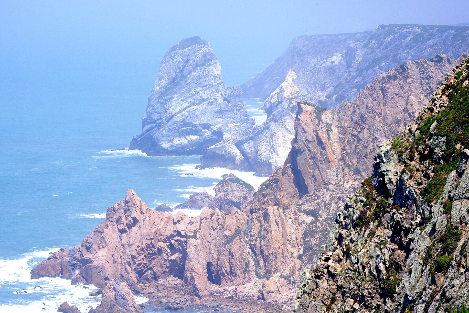 Zdjęcie przedstawia skalisty krajobraz nad morzem. Wysoki skalisty brzeg i białe fale rozbijające się o skały. Niebieskie morze zlewa się z linią horyzontu i niebem. 