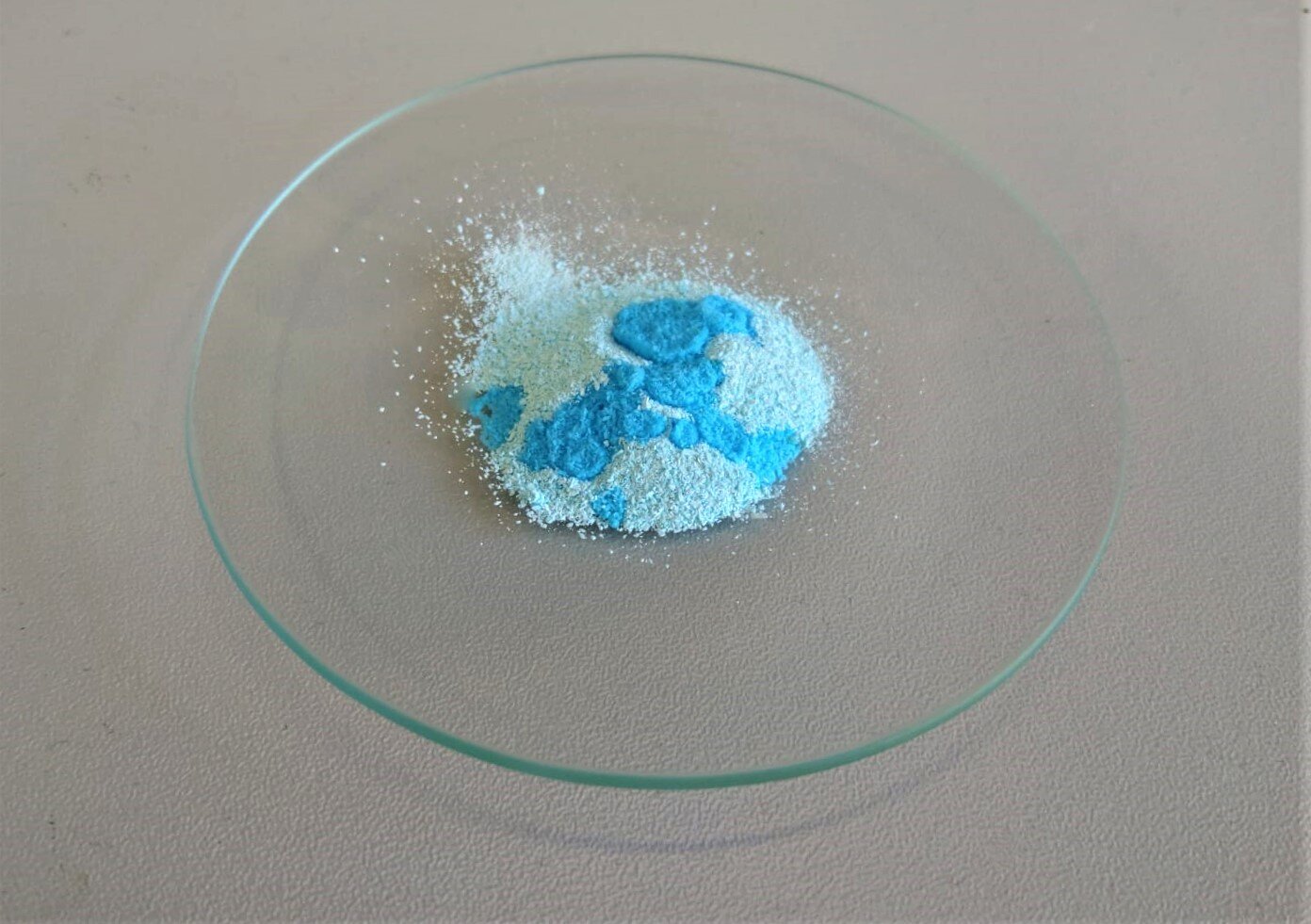 Ilustracja przedstawia szkiełko zegarkowe. Jest na nim pewna ilość jasnoniebieskiego proszku. Naniesiono na niego kilka kropli wody – w mokrych miejscach proszek jest niebieski.