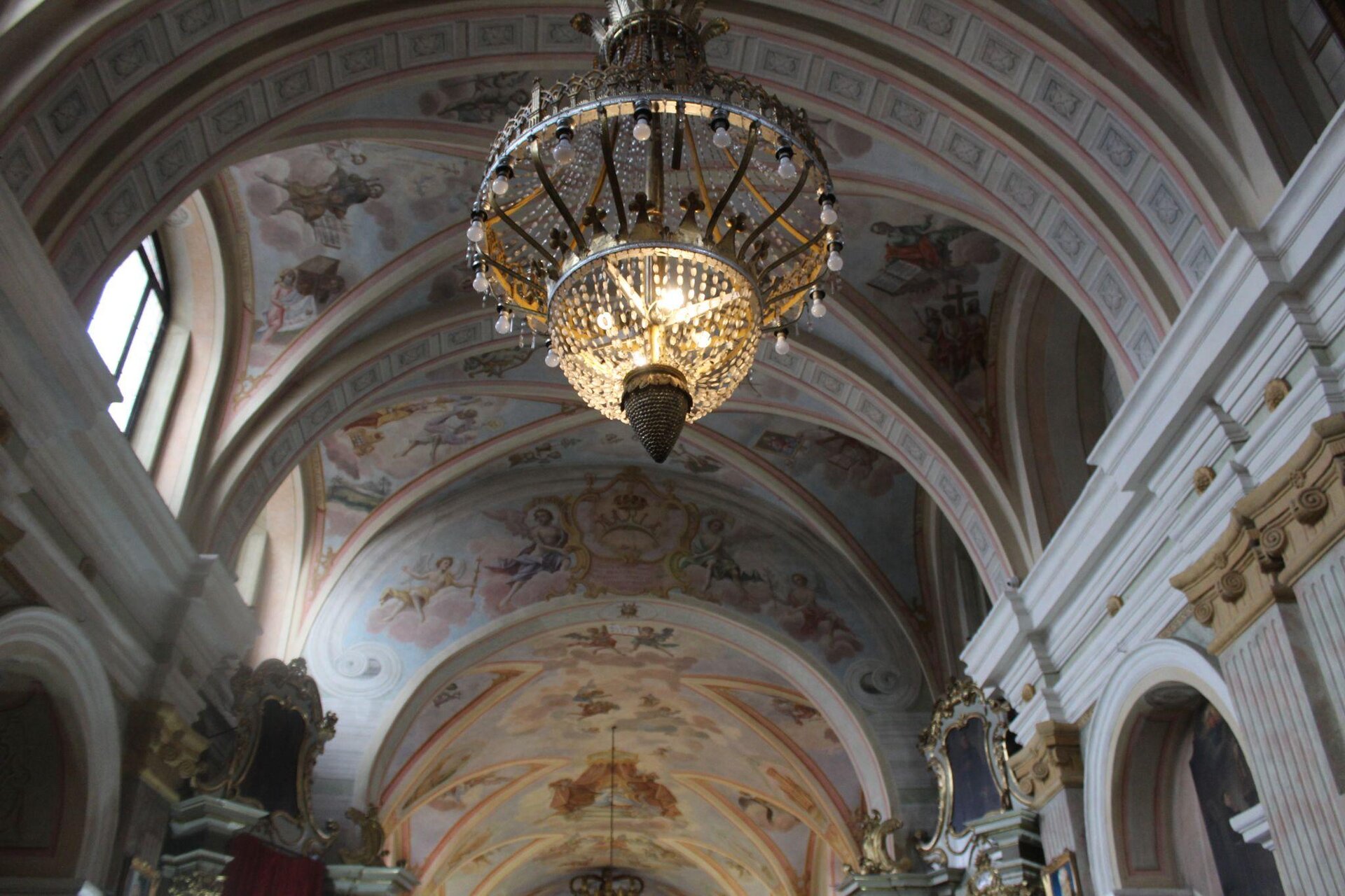 Zdjęcie przedstawia strop kościoła. Jest on bogato zdobiony obrazami, na których znajdują się sylwetki ludzkie oraz anioły. Na pierwszym planie zdjęcia widoczny jest duży, szklany żyrandol, który jest częściowo oświetlony.