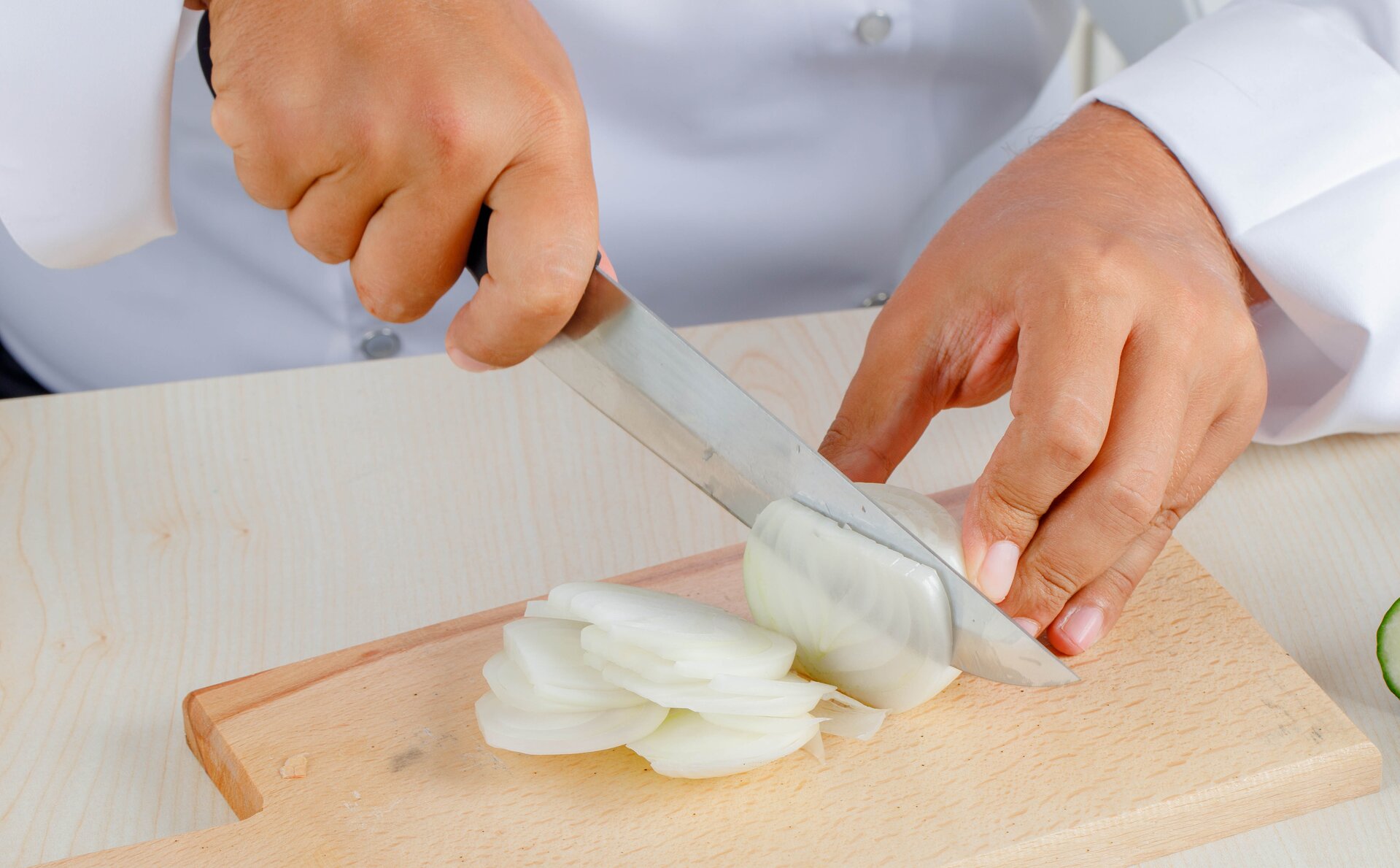 Zdjęcie przedstawia krojenie cebuli techniką emincè. Widoczna jest prostokątna drewniana deska, na której – na pierwszym planie – widoczne są cienko pokrojone plastry białej cebuli. W głębi widoczne są ręce – prawa trzyma nóż, a dłoń opiera palce na trzonku noża z metalowym ostrzem, które dotyka cebuli; lewa ręka przytrzymuje krojoną cebulę.