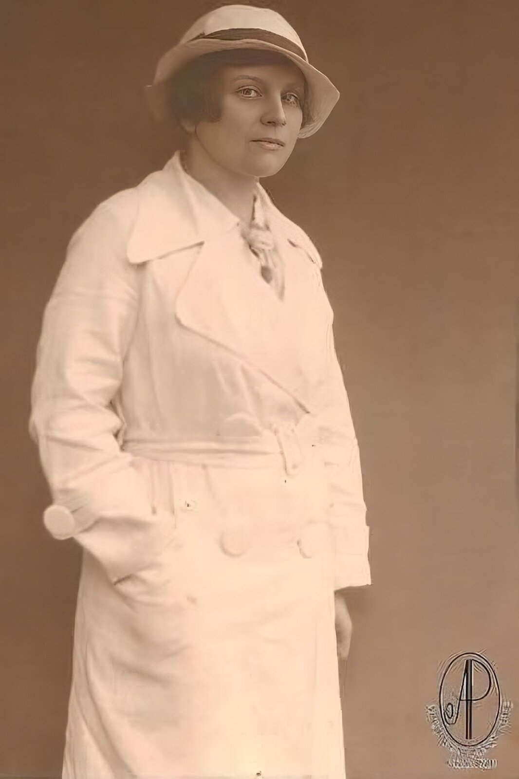 Zdjęcie przedstawia stojącą kobietę ubraną w biały, długi płaszcz. Kobieta na głowie ma biały kapelusz przewiązany czarną tasiemką. Ma krótkie, ciemne włosy. Prawą dłoń włożyła do kieszeni płaszcza, lewą trzyma swobodnie wzdłuż ciała. Kobieta pozuje na tle jednolitego tła. Zdjęcie jest sepią.