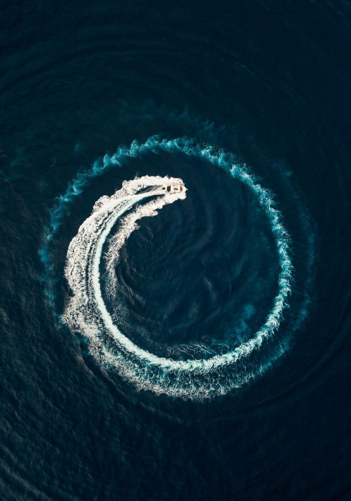 Fotografia przedstawia uchwyconą od góry ciemną powierzchnię wody, po której płynie jasna łódź. Można zauważyć trajektorię jej ruchu, śledząc ślady na wodzie – wzburzoną spiralę rozpoczynającą się z lewej strony na wysokości łodzi, rozszerzającą się w większy pierścień. Tor zaznaczony przez łódź sprawił, że woda jest jaśniejsza, biało‑niebieska. Ślady zrobione na początku są najmniej wyraźne, płynnie przechodzące w niewzburzoną powierzchnię wody.