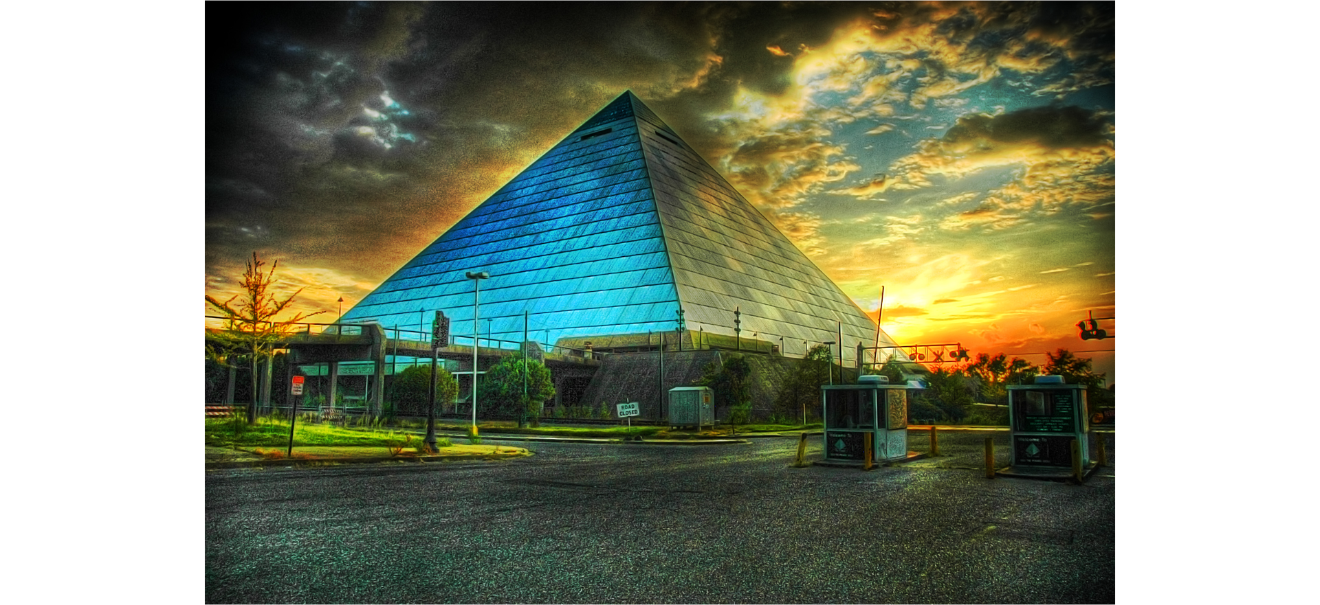 Zdjęcie przedstawia nowoczesną przeszkloną piramidę. Przed piramidą znajdują się budki parkingowe. Niebo nad piramidą jest zachmurzone.