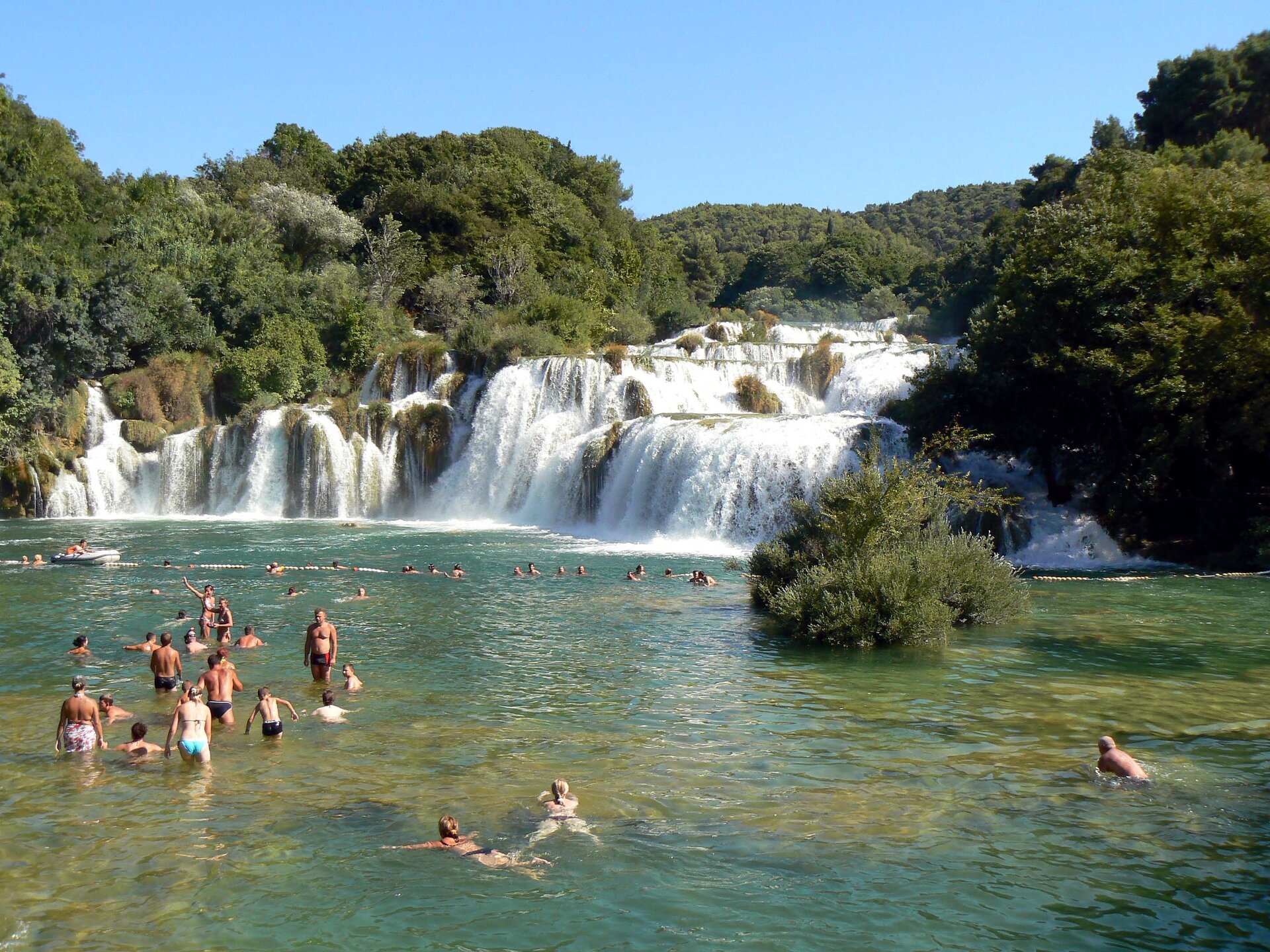 Na pierwszym planie na zdjęciu widać dwie kobiety i mężczyznę, płynących w kierunku grupy kilkunastu osób, stojących w wodzie po kolana. W oddali znajdują się ludzie kąpiący się w wodzie, która spływa po wysokich ścianach skalnych, ułożonych na kształt nieregularnych schodów. Woda spada z góry tworząc białe od piany wodospady. Po lewej stronie od grupy ludzi jest niewielka łódka, a po prawej nieduże drzewo, wystające z wody. Brzegi wodospadów porośnięte są drzewami, a w tle widać błękitne niebo.