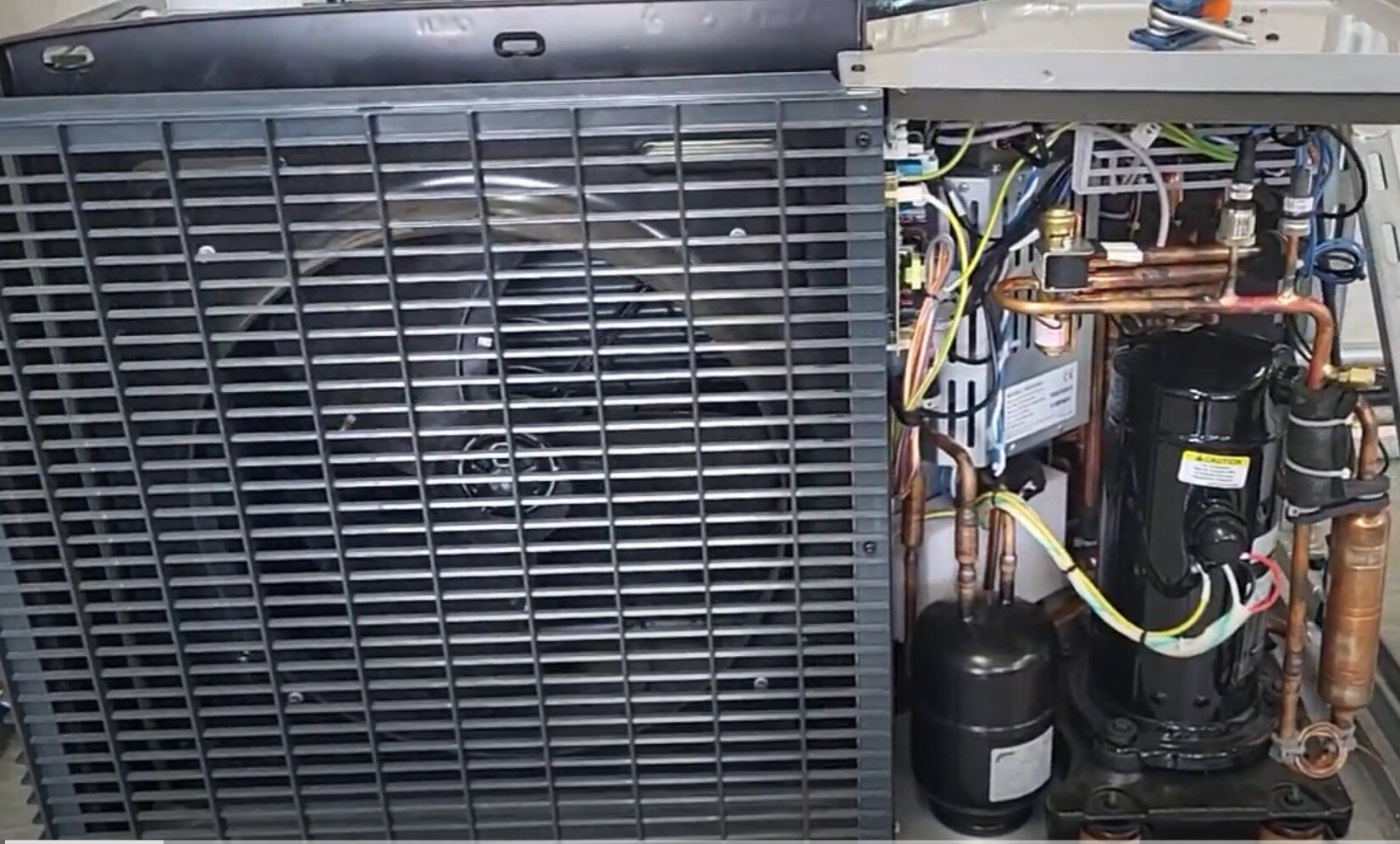 Zdjęcie ukazuje jednostkę zewnętrzną pompy typu split, widok po zdjęciu obudowy. Po lewej stronie zamiast przedniej ściany widać kratę, za którą jest widoczny wiatrak, zaś po prawej stronie widać komplet przewodów wewnętrznych jednostki.