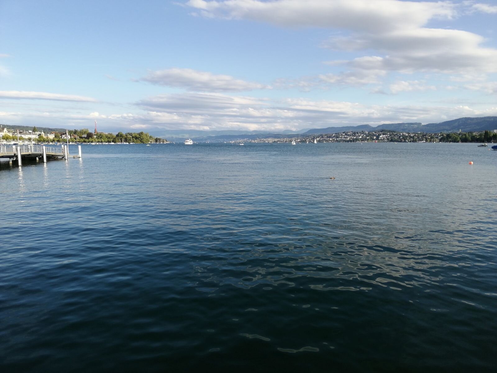 Zdjęcie przedstawia jezioro. Powierzchnia wody jest lekko falująca, daleko na horyzoncie okalają wodę zabudowania miasteczka i pasmo niewysokich gór, z boku wchodzi w zatokę koniec pomostu.  