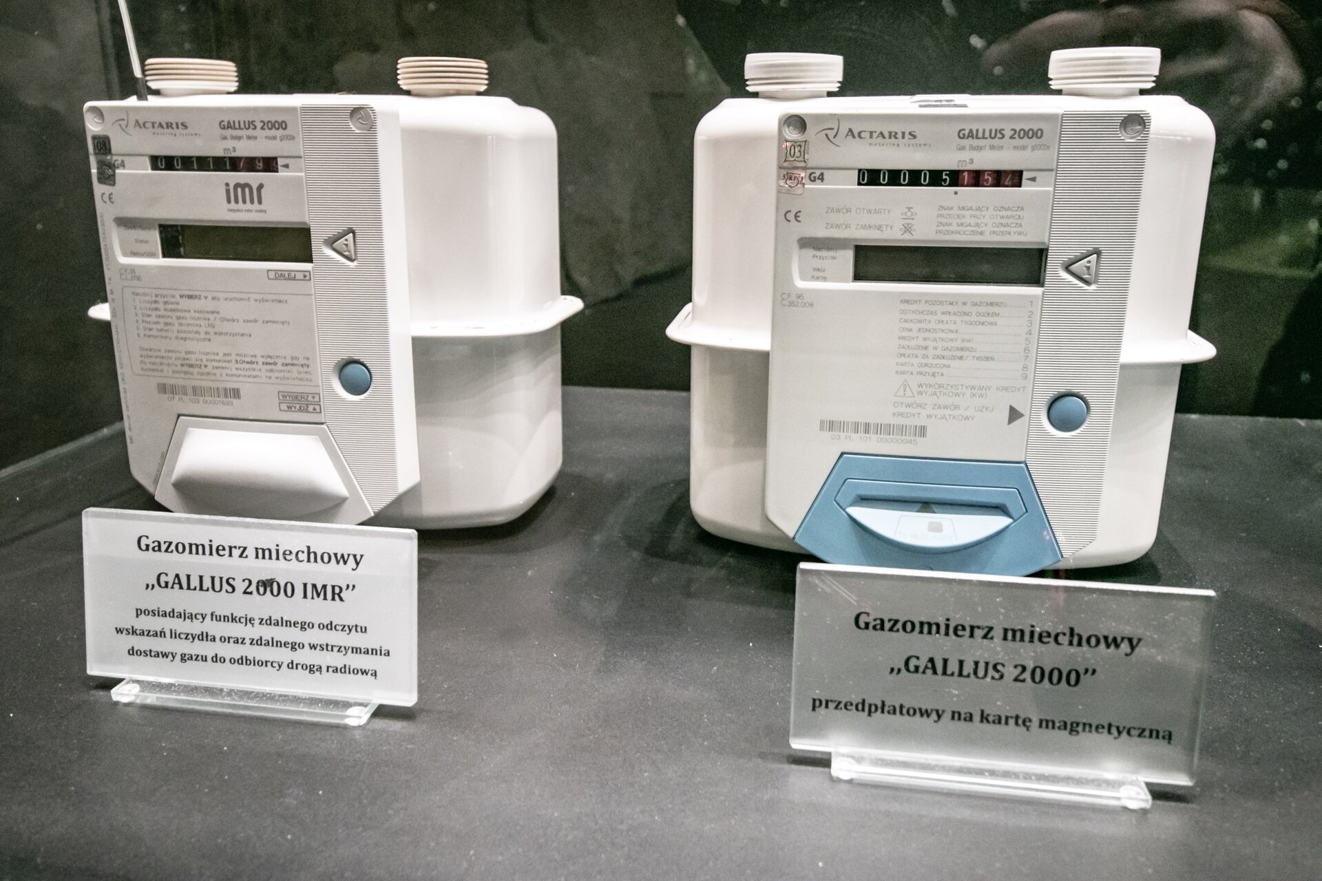 Zdjęcie przedstawia dwa gazomierze. Pierwszy z nich to GALLUS 2000 IMR. Przed gazomierzem znajduje się tabliczka z jego nazwą oraz napis: „Posiadający funkcję zdalnego odczytu wskazań liczydła oraz zdalnego wstrzymania dostawy gazu do odbiorcy drogą radiową”. Gazomierz wygląda jak pudełko z przyłączonym do jego przedniej części licznikiem. Na elemencie znajduje się zespół liczydła, wąski prostokątny ekran cyfrowy oraz w dolnej części uchwyt do ruchomego elementu. Z prawej strony znajduje się trójkątny oraz okrągły przycisk. Z górnej części pudełka wystają dwa bolce do przyłączenia przewodu gazowego.Drugi gazomierz to GALLUS 2000. Przed gazomierzem znajduje się tabliczka z jego nazwą oraz napis: „przedpłatowy na kartę magnetyczną”. Gazomierz wygląda jak pudełko z przyłączonym do jego przedniej części licznikiem. Na elemencie znajduje się zespół liczydła, wąski prostokątny ekran cyfrowy oraz w dolnej części uchwyt do ruchomego elementu. Uchwyt ma inny kolor i odrobinę inny kształt niż poprzedni gazomierz. Z prawej strony znajduje się trójkątny oraz okrągły przycisk. Z górnej części pudełka wystają dwa bolce do przyłączenia przewodu gazowego.