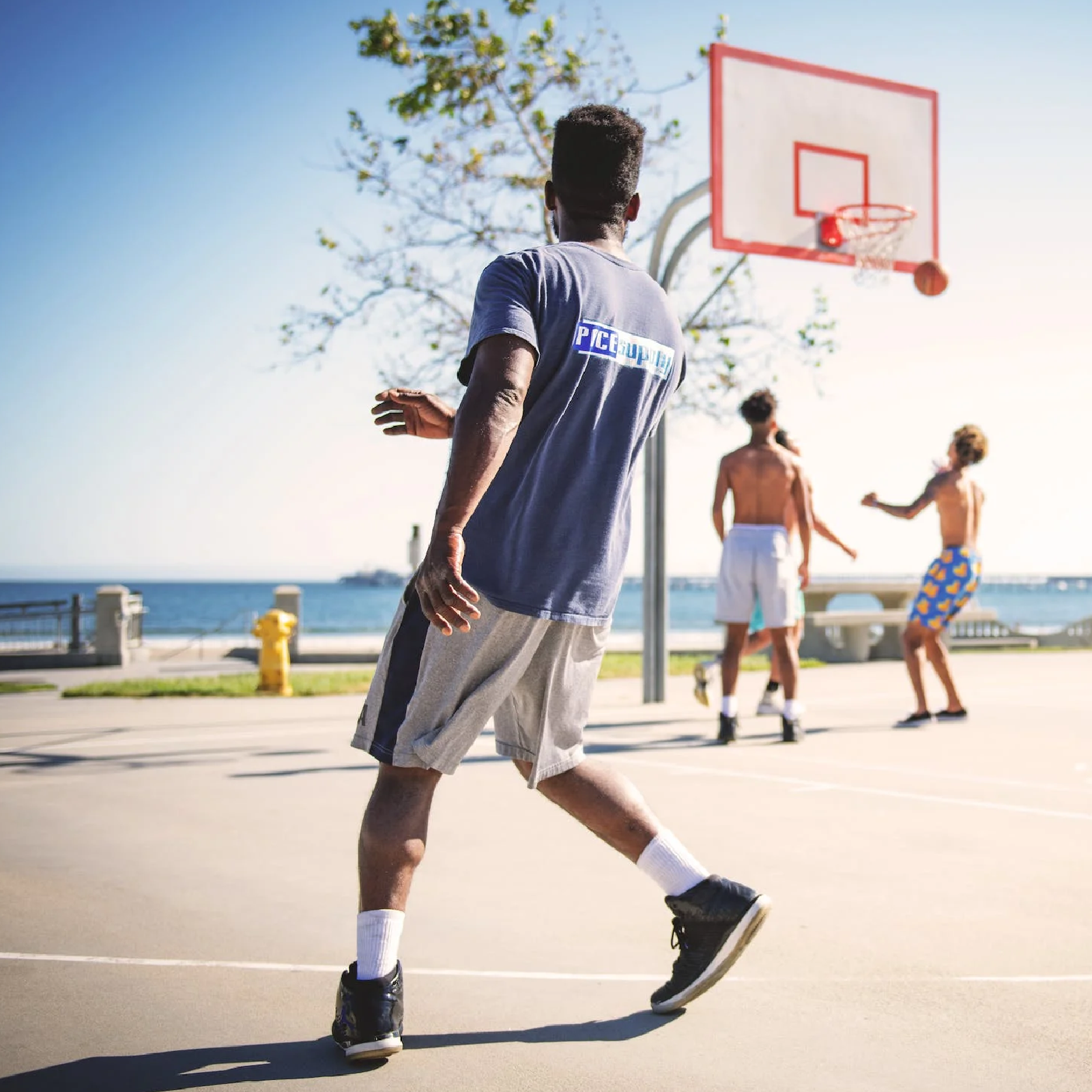 Ilustracja przedstawia czarnoskórego mężczyznę, stojącego w dużej odległości od kosza. Ubrany jest w krótkie spodenki oraz podkoszulek. Patrzy w stronę mężczyzn, grających w koszykówkę. W tle widać morze.