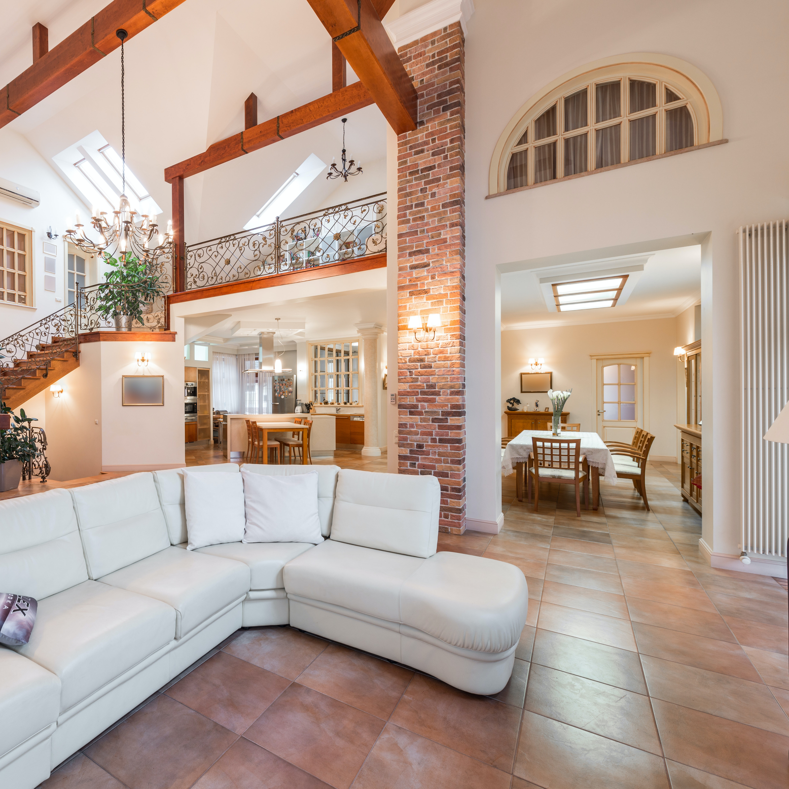 Zdjęcie przedstawia salon z antresolą, w jego centralnej części stoi duży narożnik z białej skóry. W głębi widoczny jest nakryty białym obrusem stół z krzesłami. Po lewej stronie widoczne są schody z ozdobną, metalową poręczą. Całość jest utrzymana w jasnych kolorach. 