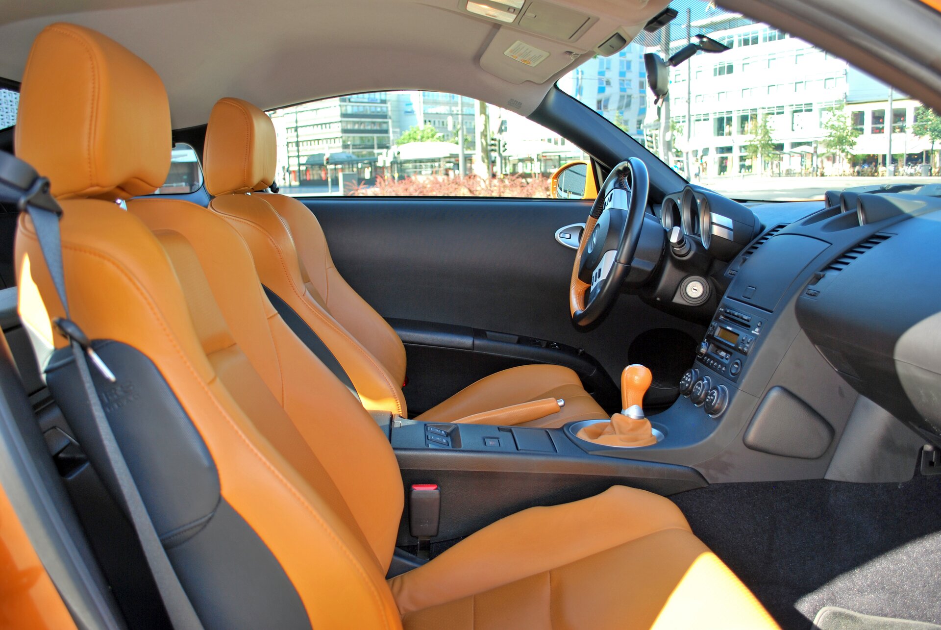 Zdjęcie przedstawia wnętrze samochodu. Ujęcie od strony pasażera na dwa pomarańczowe fotele, kierownicę i skrzynię biegów ora deskę rozdzielczą.