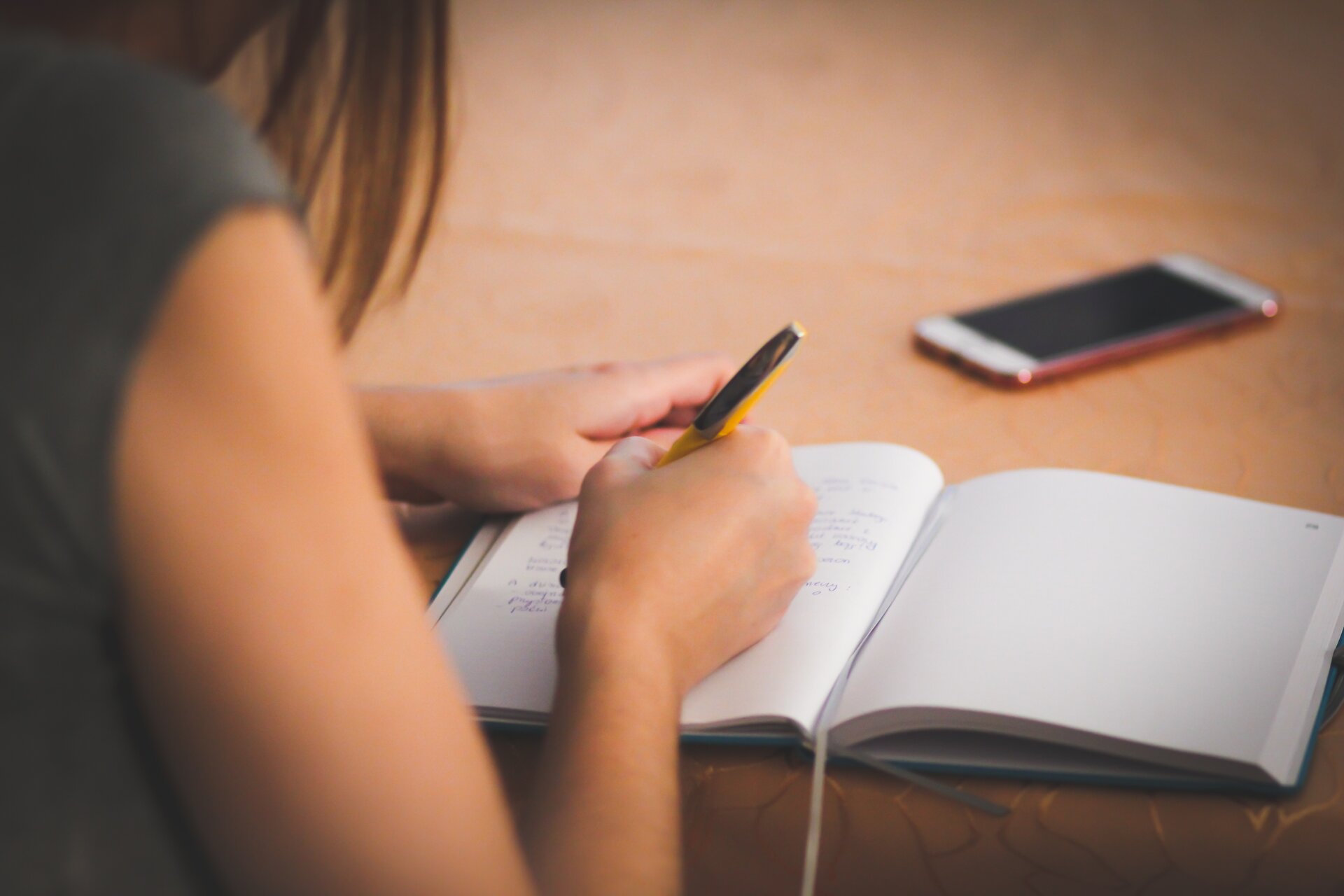 Zdjęcie przedstawia kobietę siedzącą przy biurku i piszącą w zeszycie. Obok leży telefon.