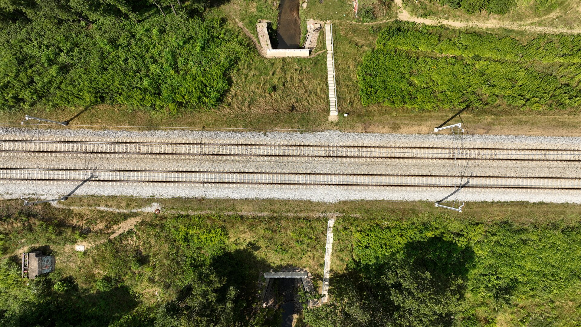 Ilustracja przedstawia przepust widoczny z lotu ptaka. Jest on niskim i wąskim tunelem wbudowanym w nasyp kolejowy, przez który przepływa strumień. Po prawej stronie zdjęcia widać schody prowadzące na szczyt nasypu. Na nasypie widoczne są dwa tory.