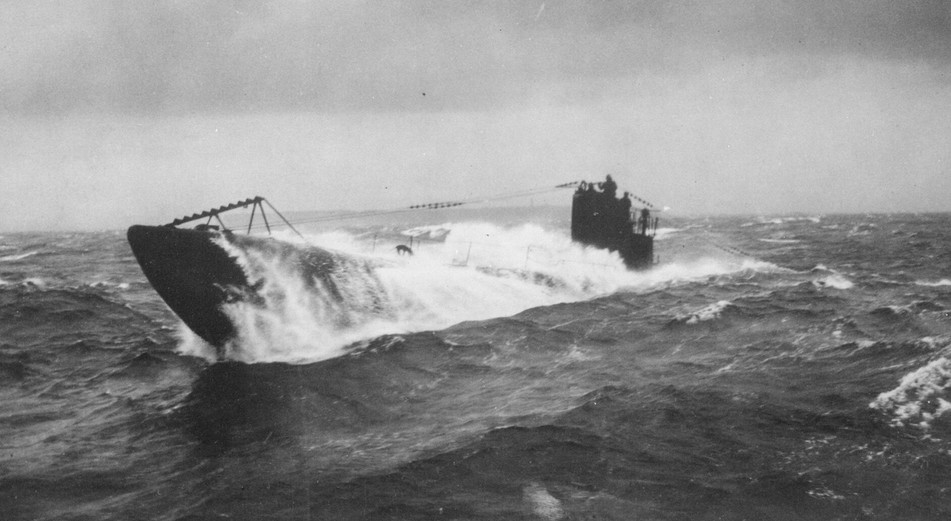 Ilustracja przedstawia niemiecki okręt podwodny wynurzający się z morza. Ma on podłużny kształt.  Widoczni są ludzie sterujący okrętem. Zdjęcie jest czarno‑białe.