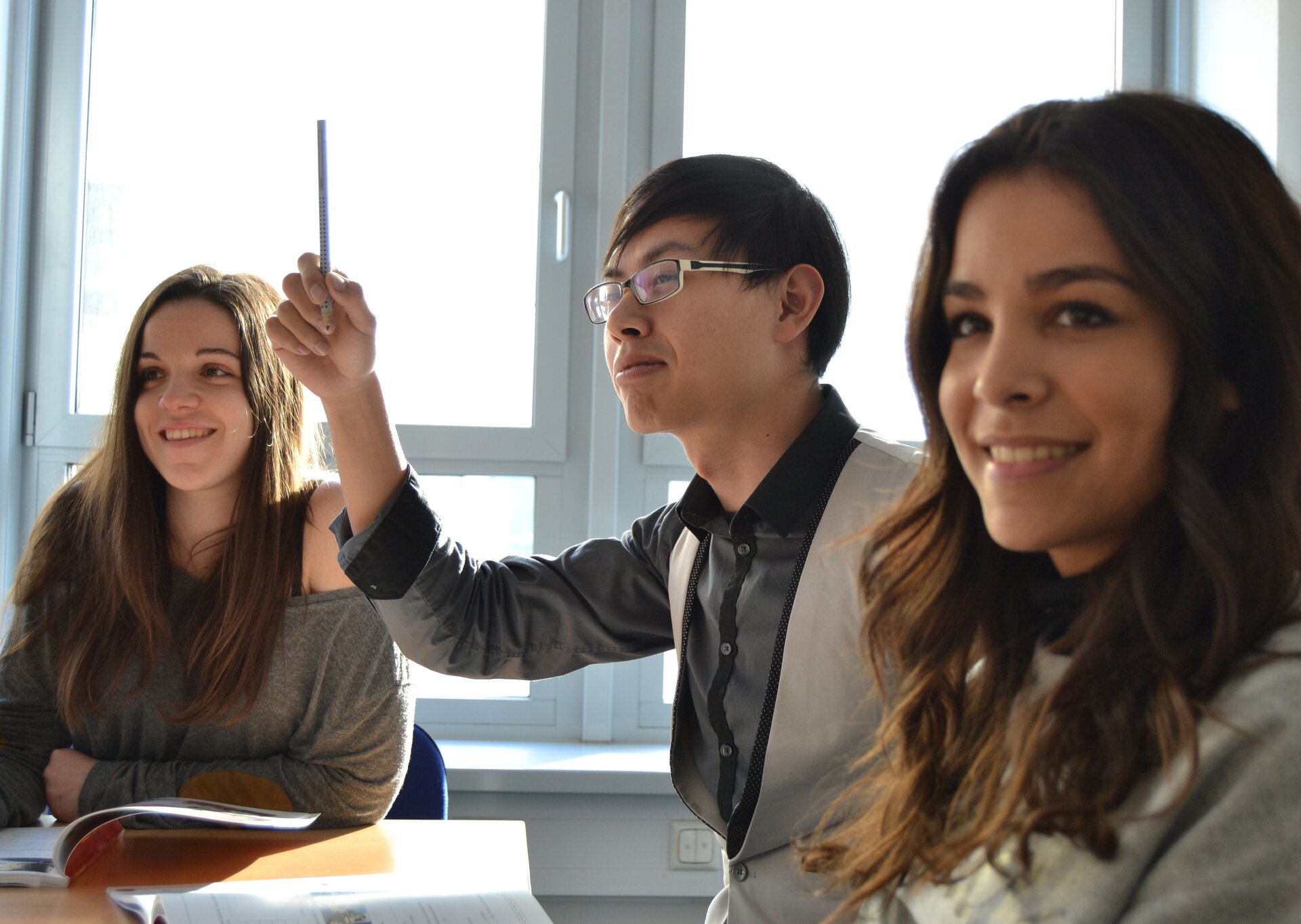 Fotografia kolorowa przedstawia spotkanie firmowe przy dużym stole. Widać dwie kobiety pomiędzy nimi siedzi mężczyzna w okularach, który podnosi rękę w której trzyma ołówek jakby zgłaszając się do odpowiedzi. A color photograph depicts a company meeting at a large table. You can see two women between them sitting a man with glasses, who raises his hand in which he is holding a pencil as if he is asking for an answer.