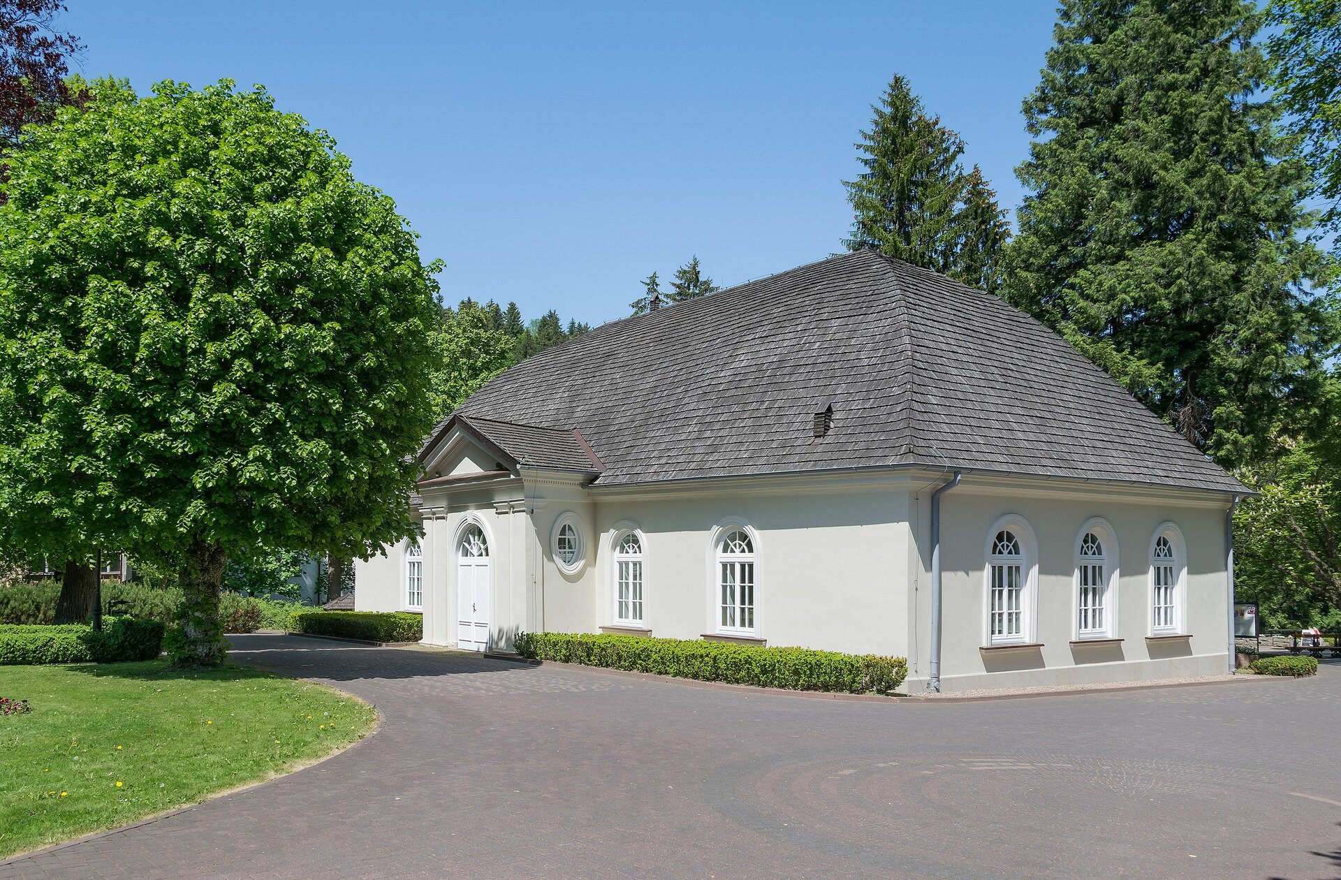 Na zdjęciu przedstawiony został jednopoziomowy, zadbany dom w białym kolorze, ze skośnym dachem o ciemnej dachówce. Okna budynku posiadają w górnej części łuki, a niektóre są całe okrągłe. Plac przed domem wyłożony jest ciemną kostką brukową. Dookoła znajduje się wiele roślinności, na przykład trawa, niski żywopłot, czy też drzewa o szerokich koronach, które porastają intensywnie zielone liście.