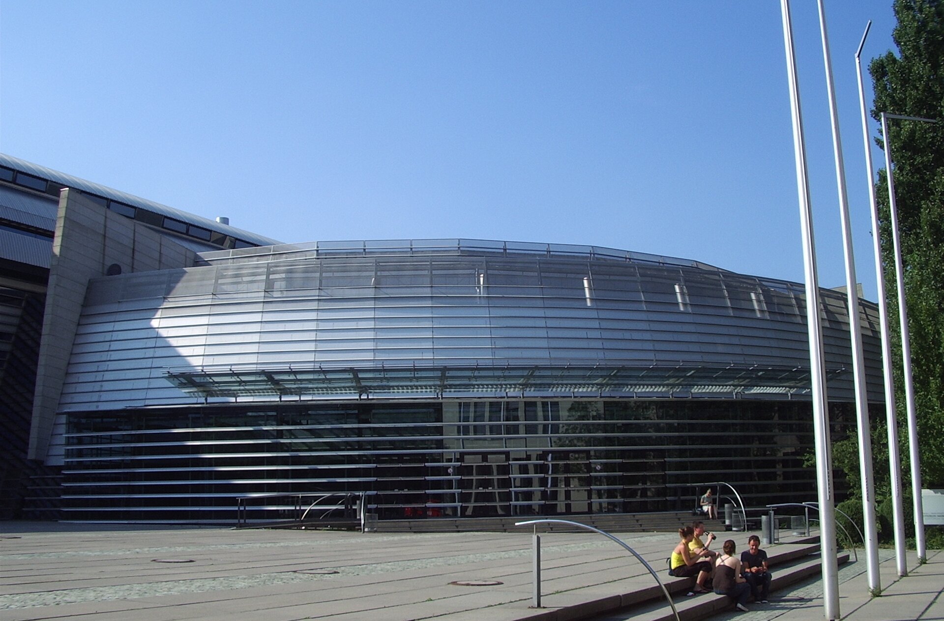 Zdjęcie przedstawia duży, nowoczesny budynek o półokrągłym kształcie. Budynek jest zbudowany ze stali i szkła. Przed budynkiem znajduje się rozległy plac.
