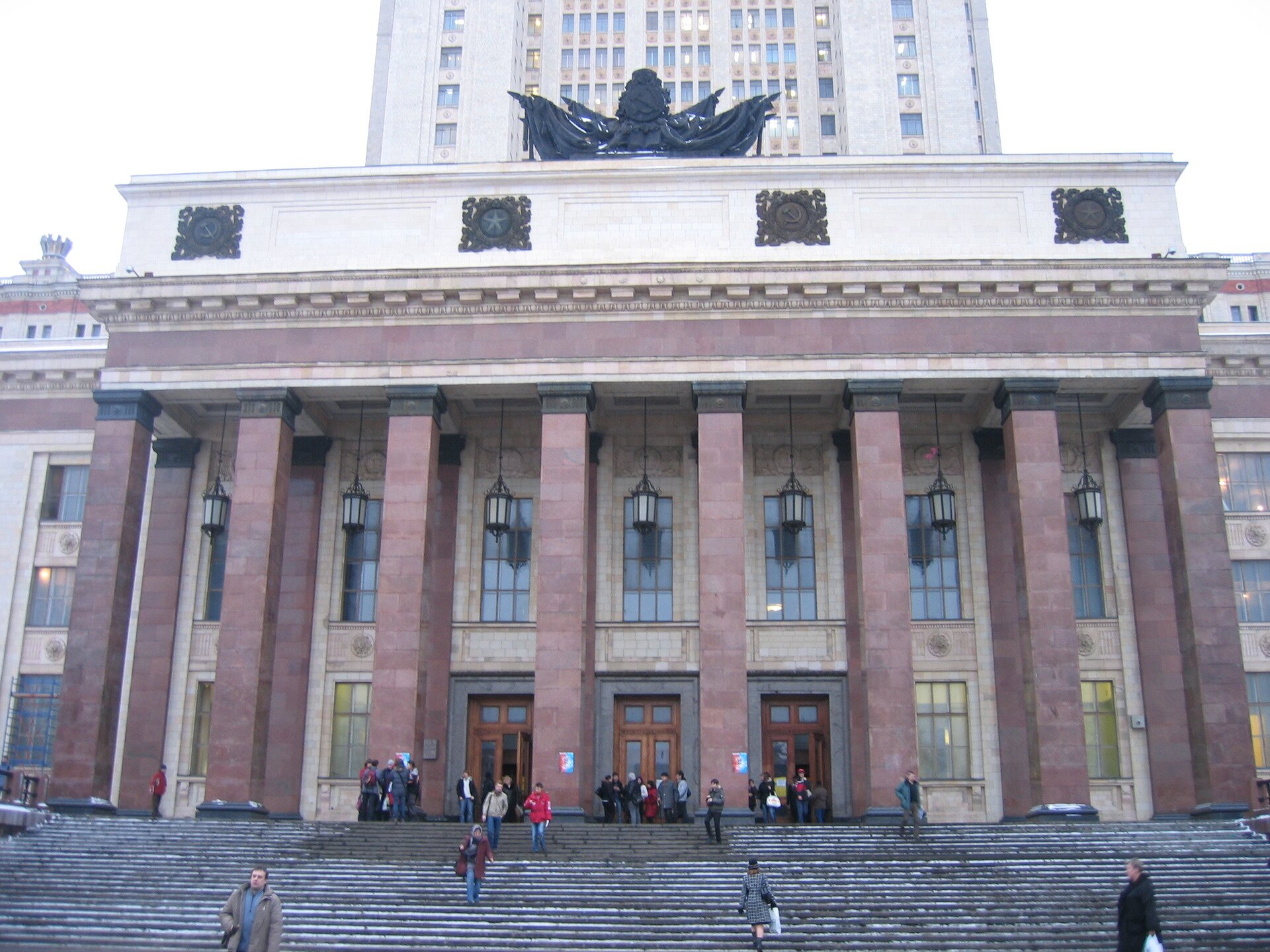 Fotografia barwna przedstawiająca budynek Uniwersytetu Moskiewskiego w Łomonosowie. Sama konstrukcja budynku opiera się na długich filarach umieszczonych na dole tuż przy schodach natomiast na szczycie budynku widać pomnik. Koło wejścia głównego gdzie widać trzy pary olbrzymich drzwi widzą pomiędzy filarami lampy.