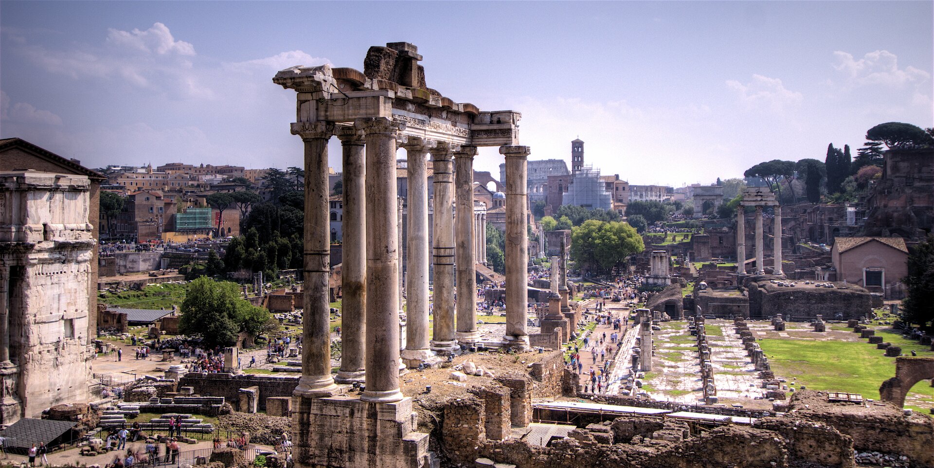 Fotografia przedstawia najstarszy plac miejski w Rzymie – Forum Romanum. Zdjęcie wykonano w słoneczny dzień, bezchmurne niebo. Na pierwszym planie w centralnej części znajdują się beżowo‑szare ruiny wysokiej kolumnady, które są pozostałościami świątyni Saturna (do współczesności zachowało się osiem kolumn). Po obu stronach kolumnady na ziemi są fragmenty kamieni i pozostałości dawnych zabudowań. Na drugim planie – porośnięte jasnozieloną trawą tereny, fragmenty ruin odgrodzone od zwiedzających metalowymi barierkami, po lewej stronie duża, zwarta bryła architektoniczna, która jest bokiem łuku triumfalnego. Na chodnikach wokół centralnie umieszczonej kolumnady znajduje się wiele idących osób. Nieco dalej, po prawej stronie jest mniejsza kolumnada, złożona z trzech kolumn. Charakterystyczna dla Rzymu roślinność, malownicze drzewa i krzewy, a także budynki, zza których w oddali wyłania się fragment Koloseum.