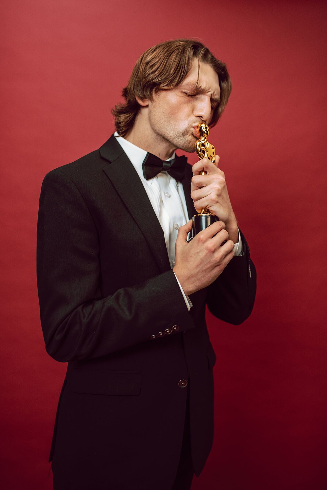 Zdjęcie przedstawia mężczyznę w średnim wieku ubranego w czarny garnitur, białą koszulę ozdobioną czarną muchą pod szyją.  Mężczyzna ma w dłoniach statuetkę Oskara, którą całuje w głowę. 