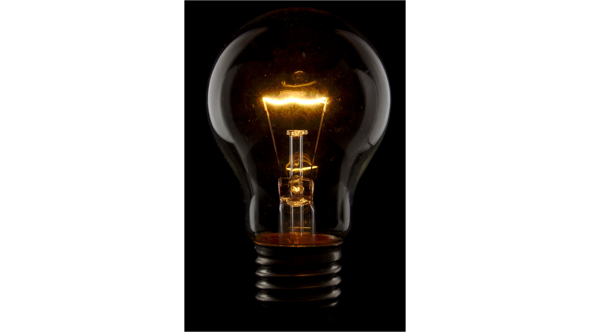 Zdjęcie przedstawia świecą, klasyczną żarówkę na czarnym tle. Bańka żarówki jest przezroczysta, żarnik świeci żółtym światłem. Tło ciemne.