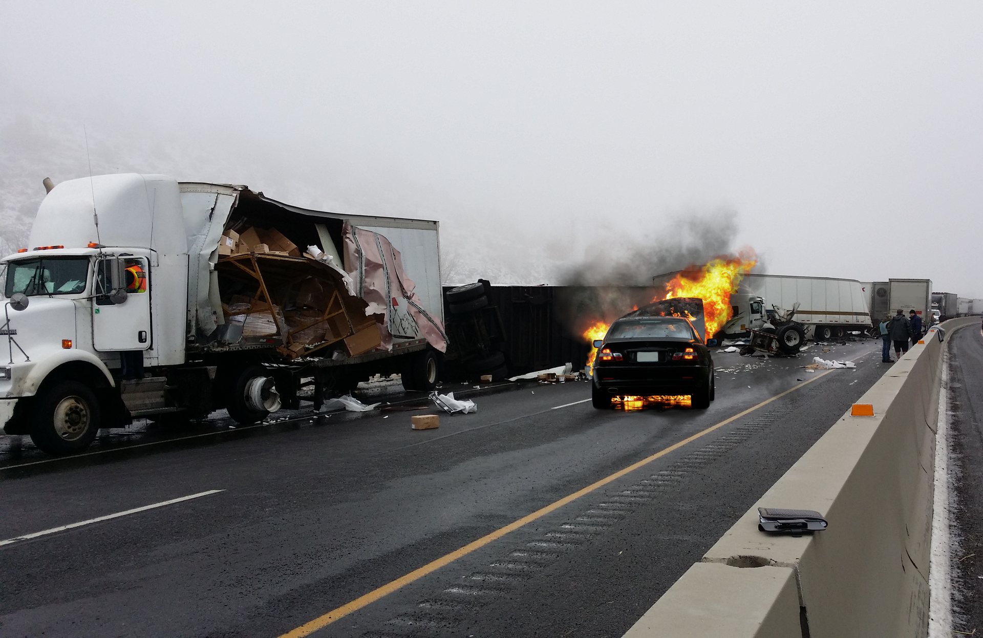 Dzień. Dwupasmowa jezdnia. Wzdłuż lewego pasa jezdni samochody ciężarowe stojące jeden za drugim. Po prawej duża biała ciężarówka. Bok naczepy całkowicie rozerwany. Wewnątrz naczepy zniszczone przedmioty. Na jezdni kilka porozrzucanych rzeczy. W głębi zdjęcia, za ciężarówki, dwie duże ciężarówki również uszkodzone. Na prawym pasie jezdni, samochód osobowy. Przód samochodu skierowany w stronę ciężarówek w głębi zdjęcia. Maska samochodu otwarta. Przód samochodu pali się. Po prawej stronie, betonowy mur wzdłuż krawędzi jezdni. Na murze notes.