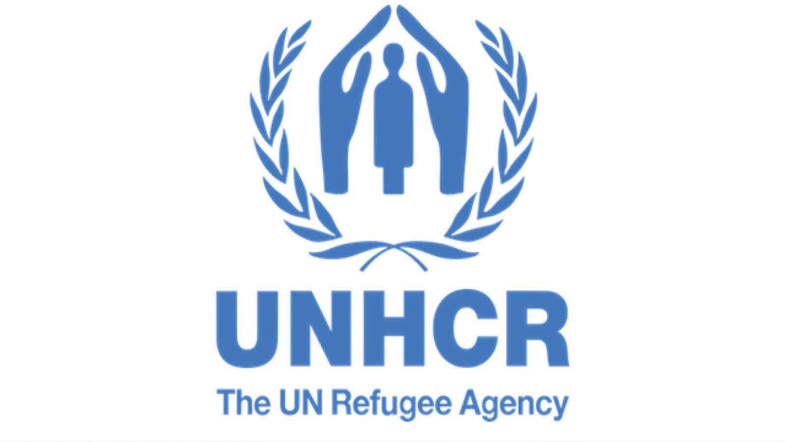 Grafika przedstawia logo w kolorze niebieskim na białym tle. U góry widoczna jest para dłoni tworząca schronienie nad postacią człowieka, otoczona gałązkami oliwnymi symbolizującymi pokój. Poniżej widoczny jest napis w języku angielskim: UNHCR The UN Refugee Agency.