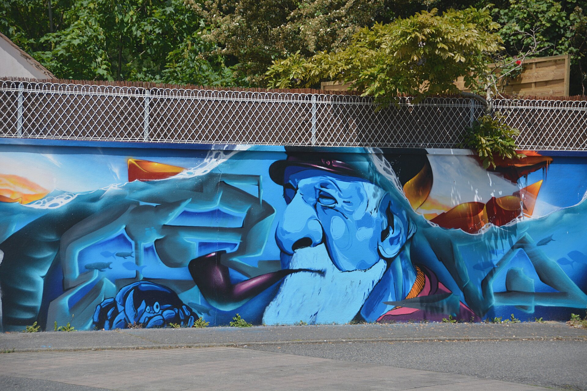 Zdjęcie przedstawia mural wykonany na betonowym ogrodzeniu miejskim. To twarz starego mężczyzny z fajką.
