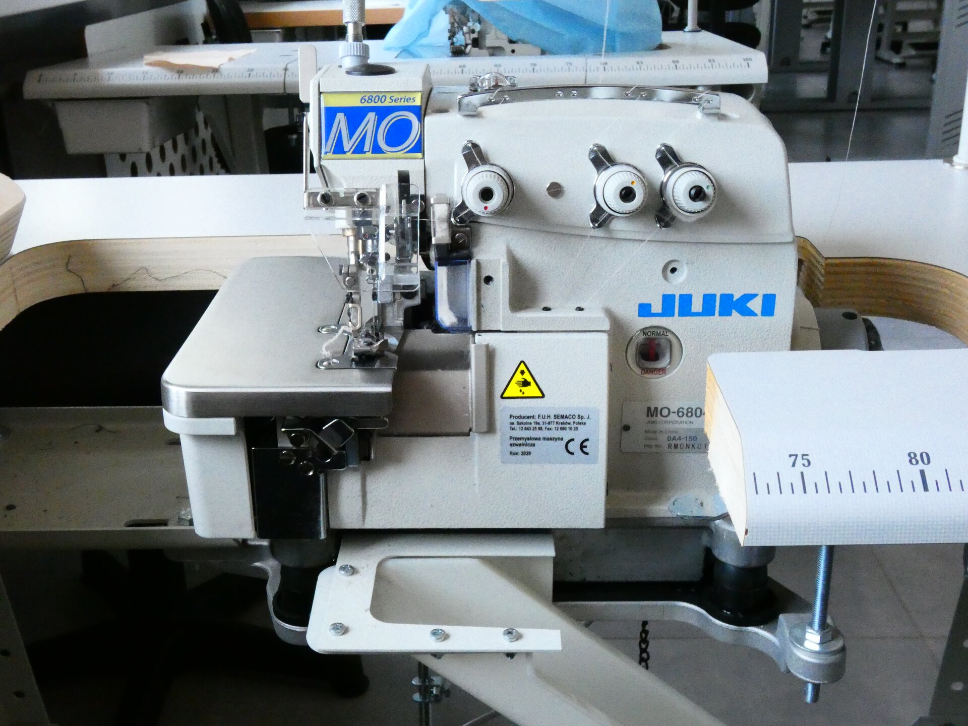 Zdjęcie przedstawia białą maszynę. Z lewej strony ma metalową tackę, jest otoczona deską z centymetrową podziałką. Z przodu znajdują się trzy pokrętła.