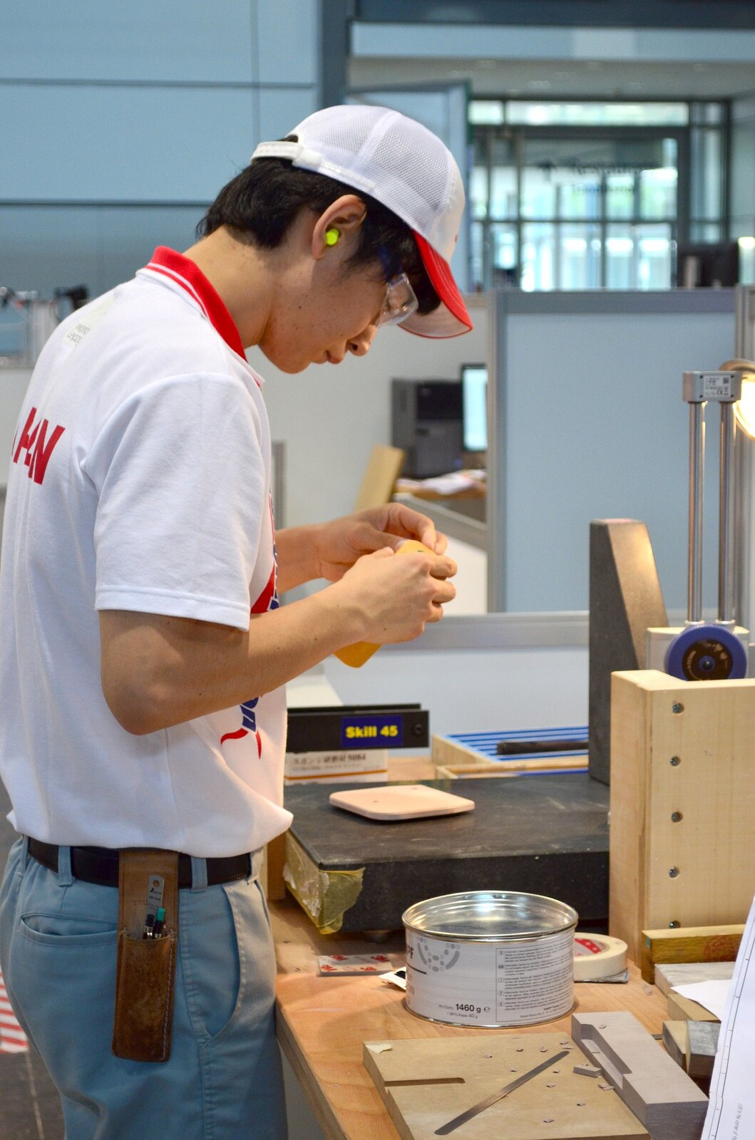 kliknij aby powiększyć Na zdjęciu widać młodego człowieka pracującego w drewnie. Ubrany w białą koszulkę z czerwonymi napisami.
Na głowie biała czapka z czerwonym daszkiem. W uszach stopery, na oczach okulary ochronne.
Przy pasku od spodni ma doczepiony piórnik, a nim ołówki. Stoi przy stole, na którym znajdują się niezbędne materiały w jego pracy.
