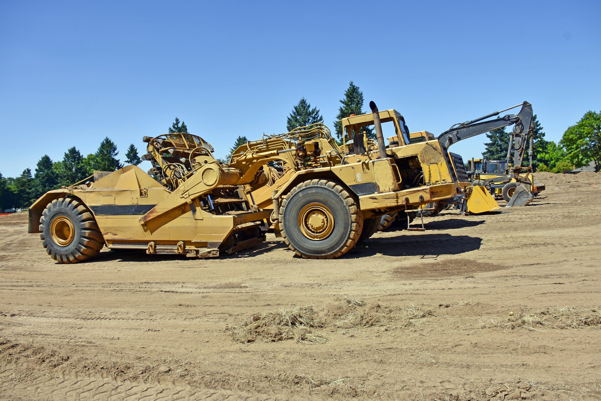 Zdjęcie przedstawia zgarniarkę. Widoczna jest maszyna w ujęciu z boku, stojąca na rozjeżdżonej i ubitej ziemi. W środkowej części widoczna jest rama, czyli konstrukcja nośna, osadzona w przegubie ciągnika i w czopach skrzyni. Z tyłu widoczna jest skrzynia (pojemnik), zaś na przodzie układ ładowania oraz kabina operatora. Maszyna jest bardzo rozbudowana.

Opisy obiektów prezentowanych na grafikach są załączone w treści galerii zdjęć.
