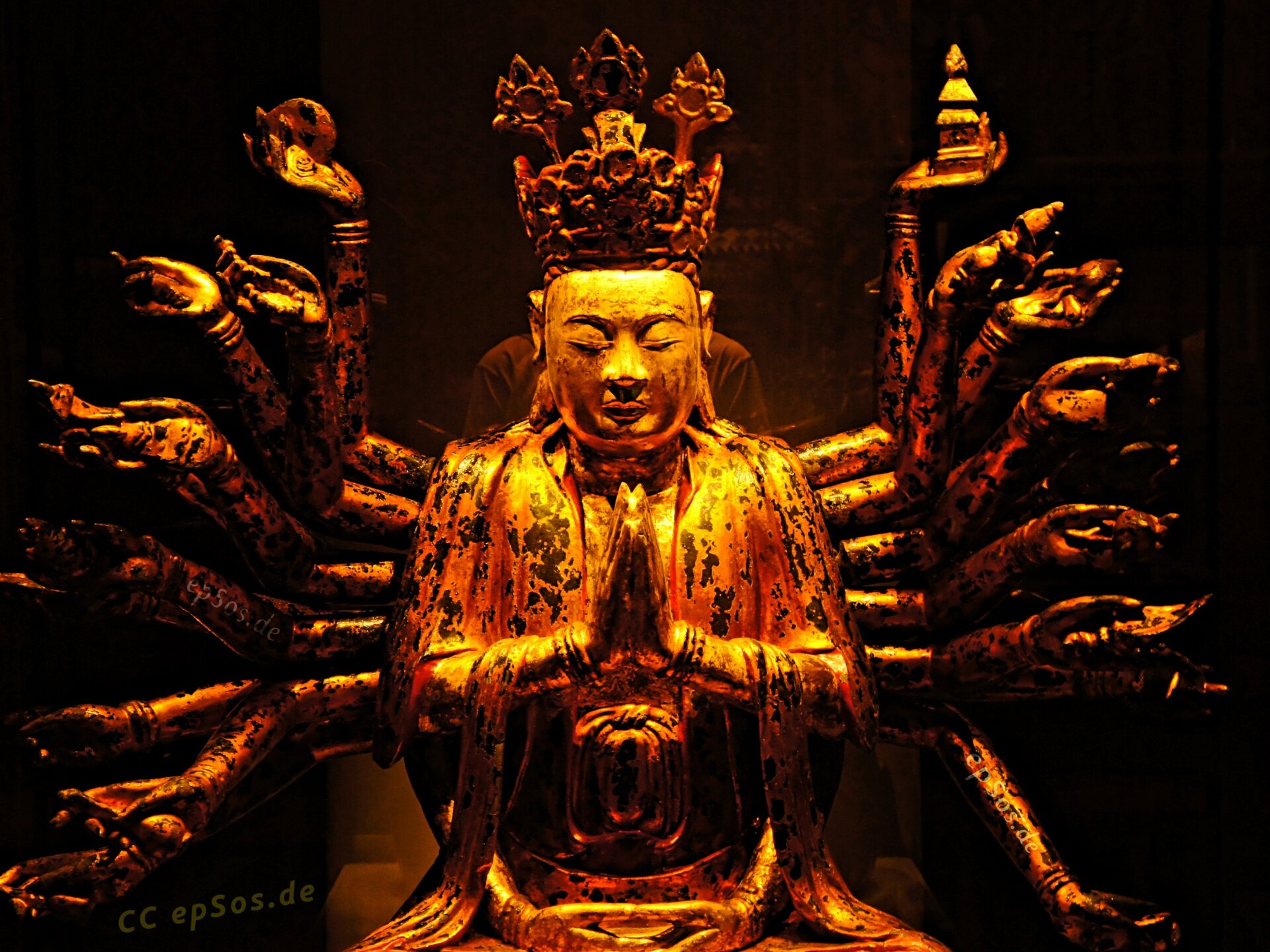 Fotografia posągu Buddy. Ma zamknięte oczy, ręce składa w geście modlitwy. Na głowie ma koronę. Za nim znajduje się wiele par rąk, które układają się wokół niego jak promienie słońca.